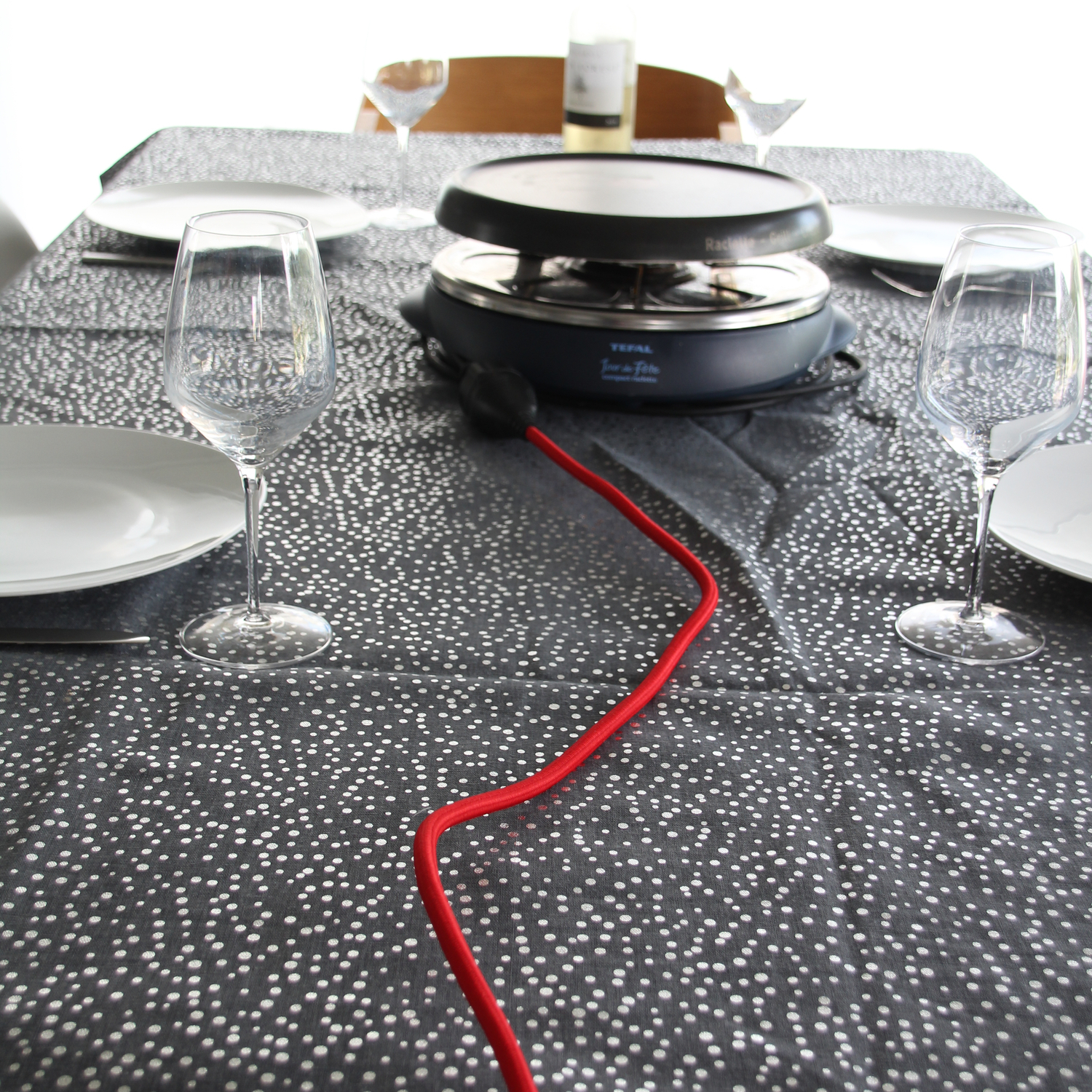 Table avec grill à raclette, assiettes, verres à vin et nappe à pois.