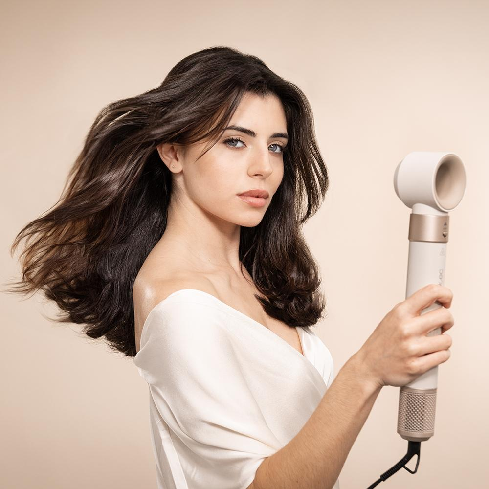 Una mujer sostiene un secador de pelo, con el pelo suelto. El fondo es de un color suave y neutro.