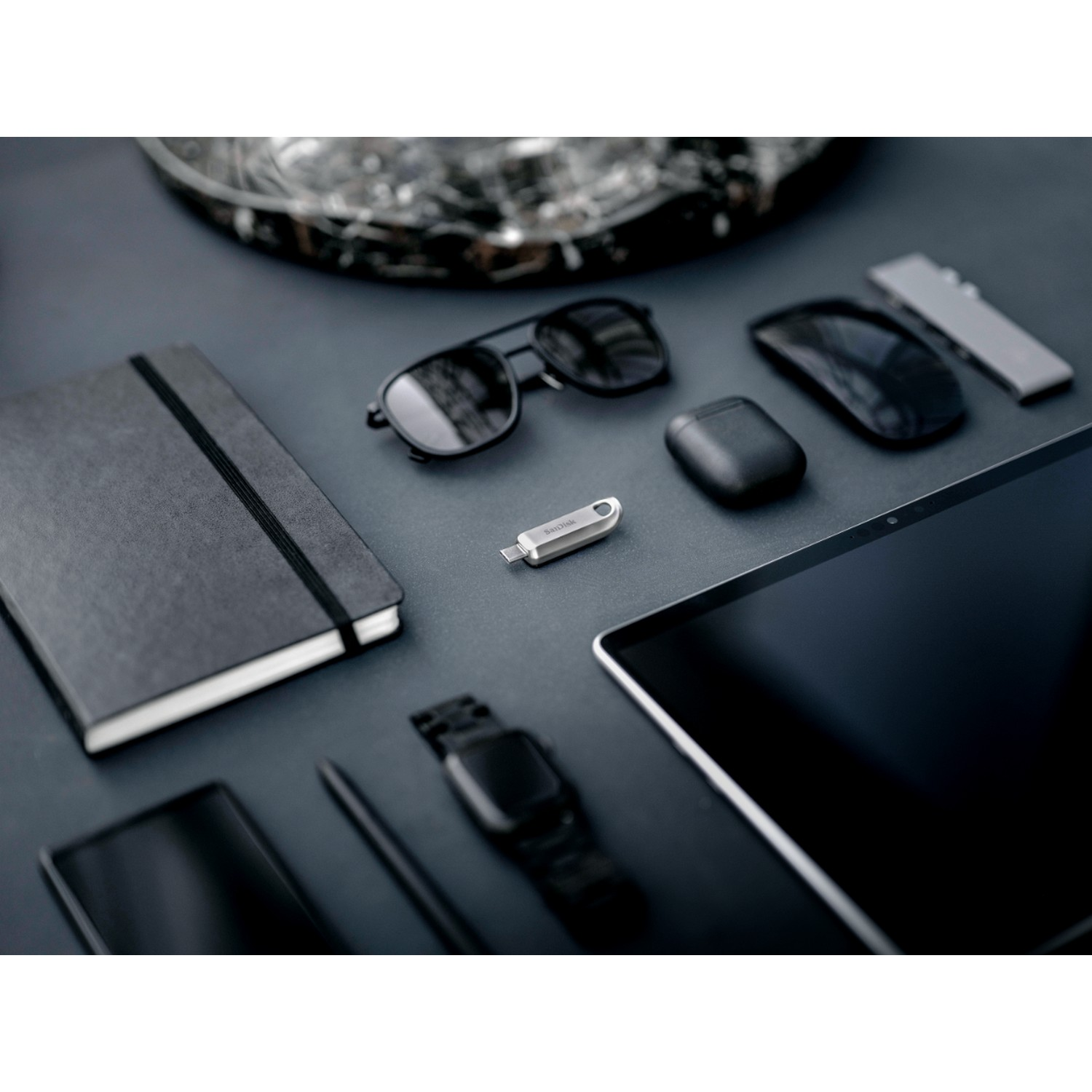 Surtido de artículos tecnológicos: unidad USB-C, bolígrafo, gafas de sol, tableta, auriculares, reloj, todo oscuro.