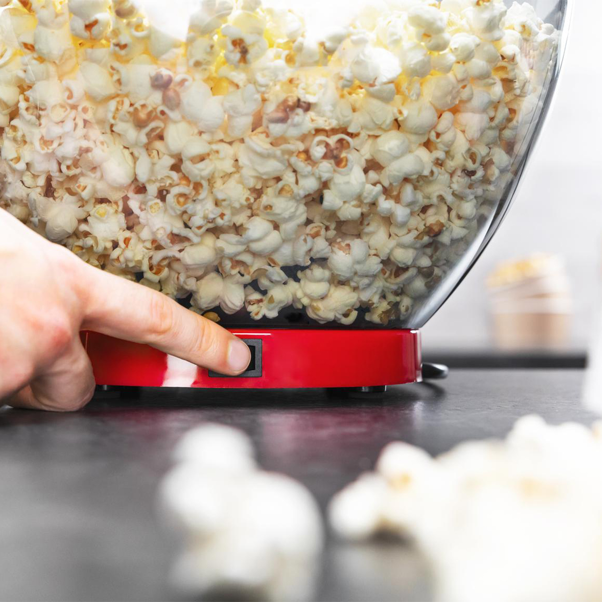 Una mano presiona un botón en una máquina de palomitas roja llena de palomitas, lista para servir, sobre negro.