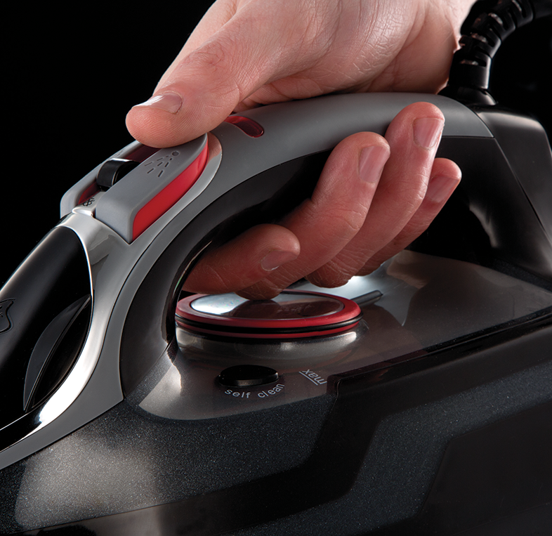 Kéz egy szürke és fekete vasalót tart, egy gombot nyom meg a tetején.