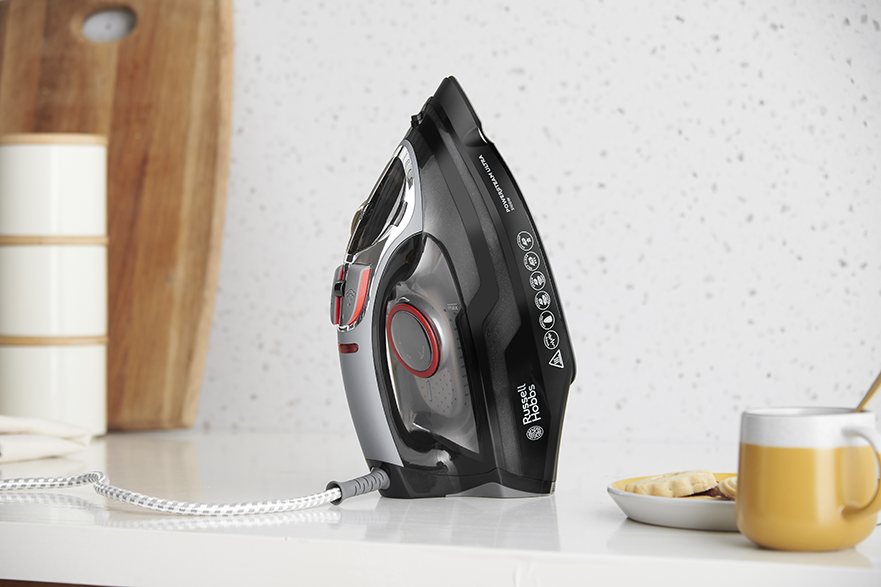 Egy vasaló fehér felületen, fa deszkával és egy bögrével a háttérben.