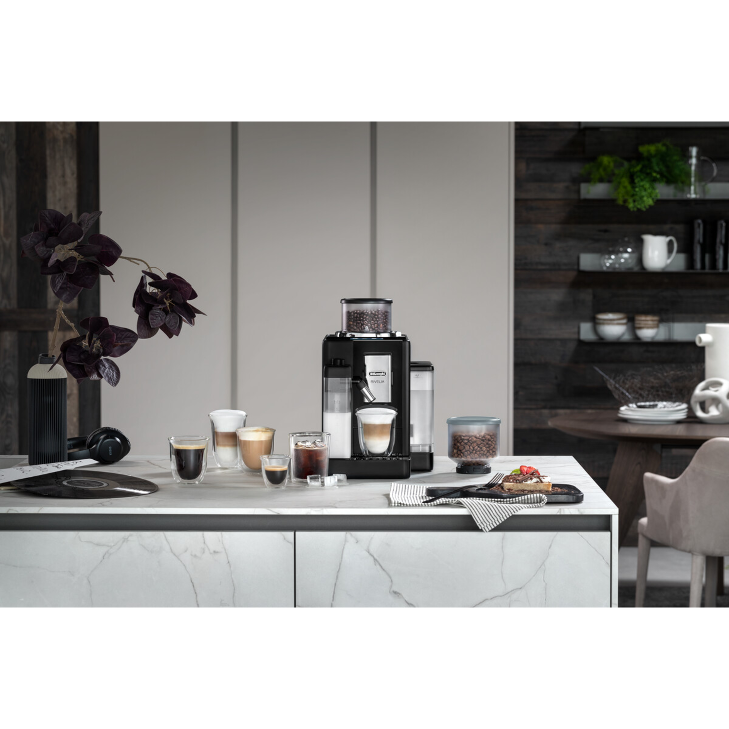 Coffee machine with glasses of coffee, flowers, and a dessert on a countertop. Wooden background.