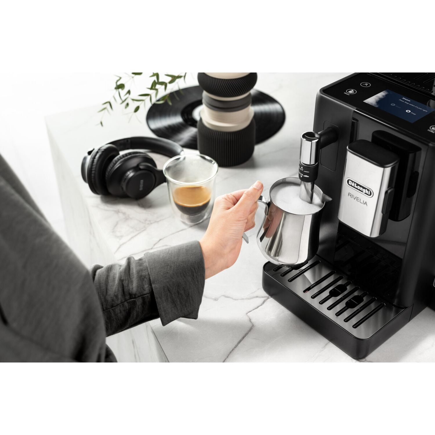 Een persoon giet melk in een koffiemachine, met een glas espresso en een koptelefoon in de buurt.