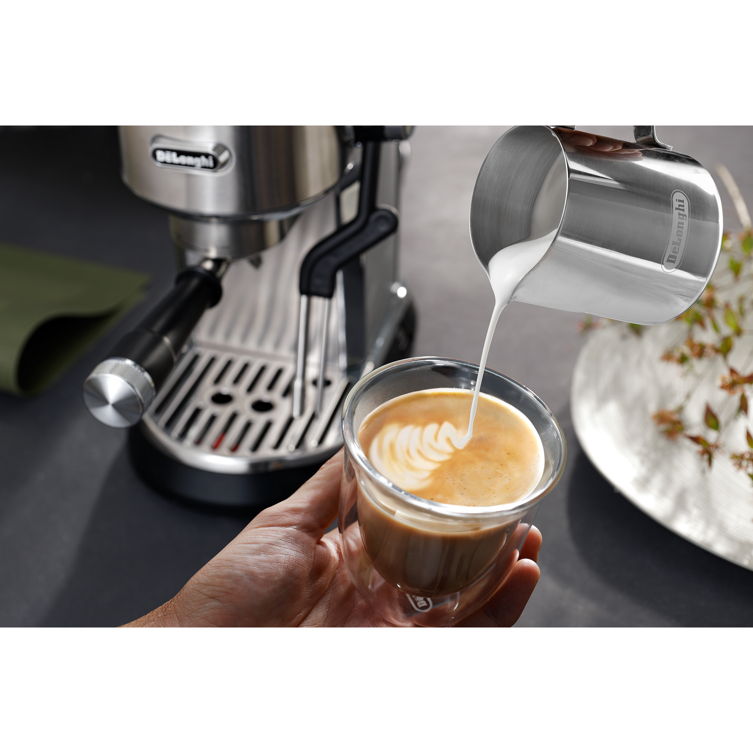 A person pouring milk from a stainless steel pitcher into a glass of coffee. A coffee machine is visible in the background.