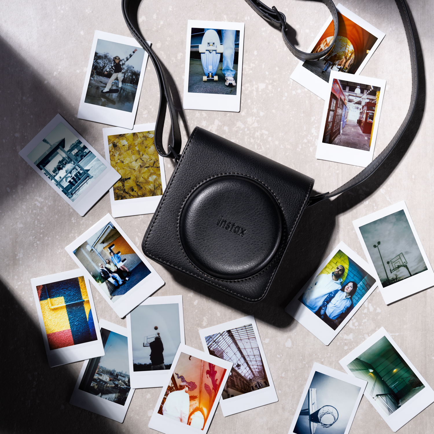 Een zwarte Instax camerahoes met Instax foto's eromheen, op een lichtgrijze ondergrond. Een riem is zichtbaar.