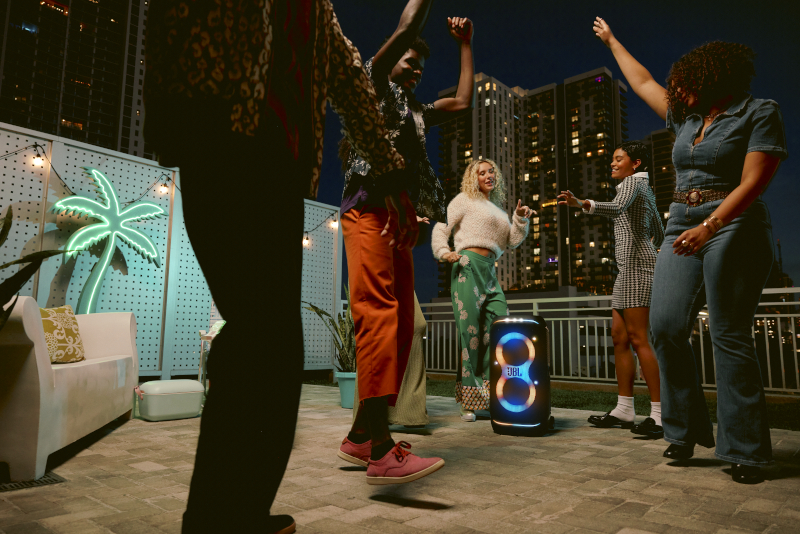 Grupa ludzi tańczących na dachu, z głośnikiem i światłami miasta.