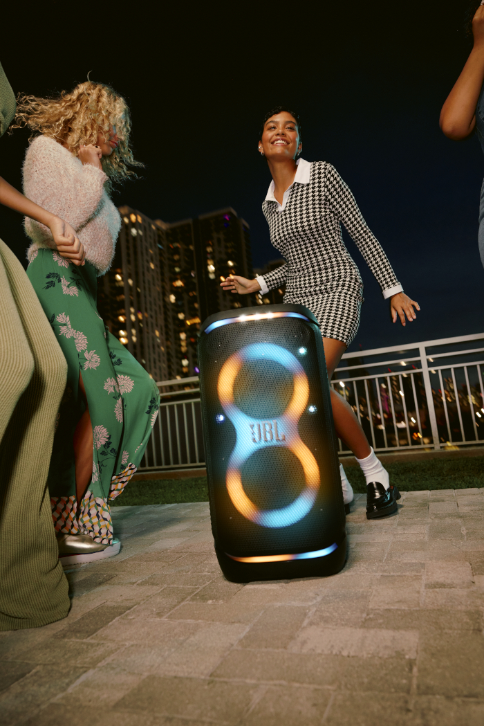 Ludzie tańczą wokół głośnika JBL w nocy, światła miasta w tle.