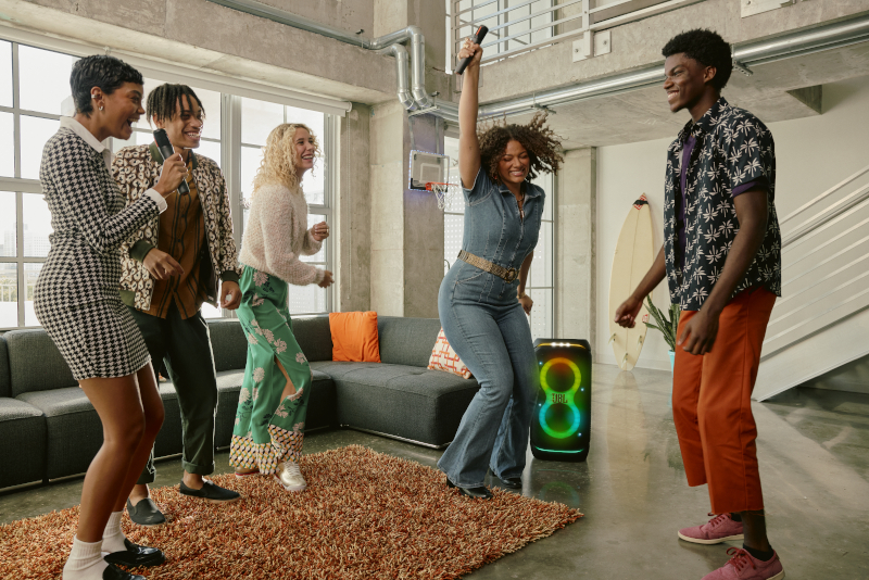 Grupa ludzi śpiewających karaoke. Kobieta z mikrofonem śpiewa. Głośnik jest po prawej stronie. Pokój z meblami.