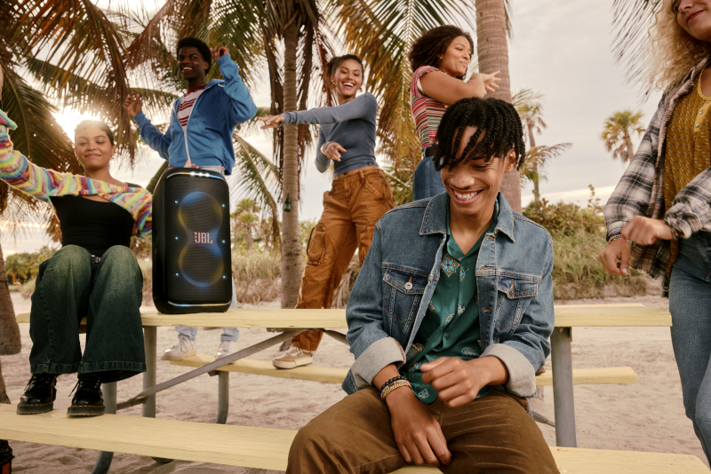 Grupa ludzi tańczy obok czarnego głośnika JBL na piaszczystej plaży, palmy w tle.