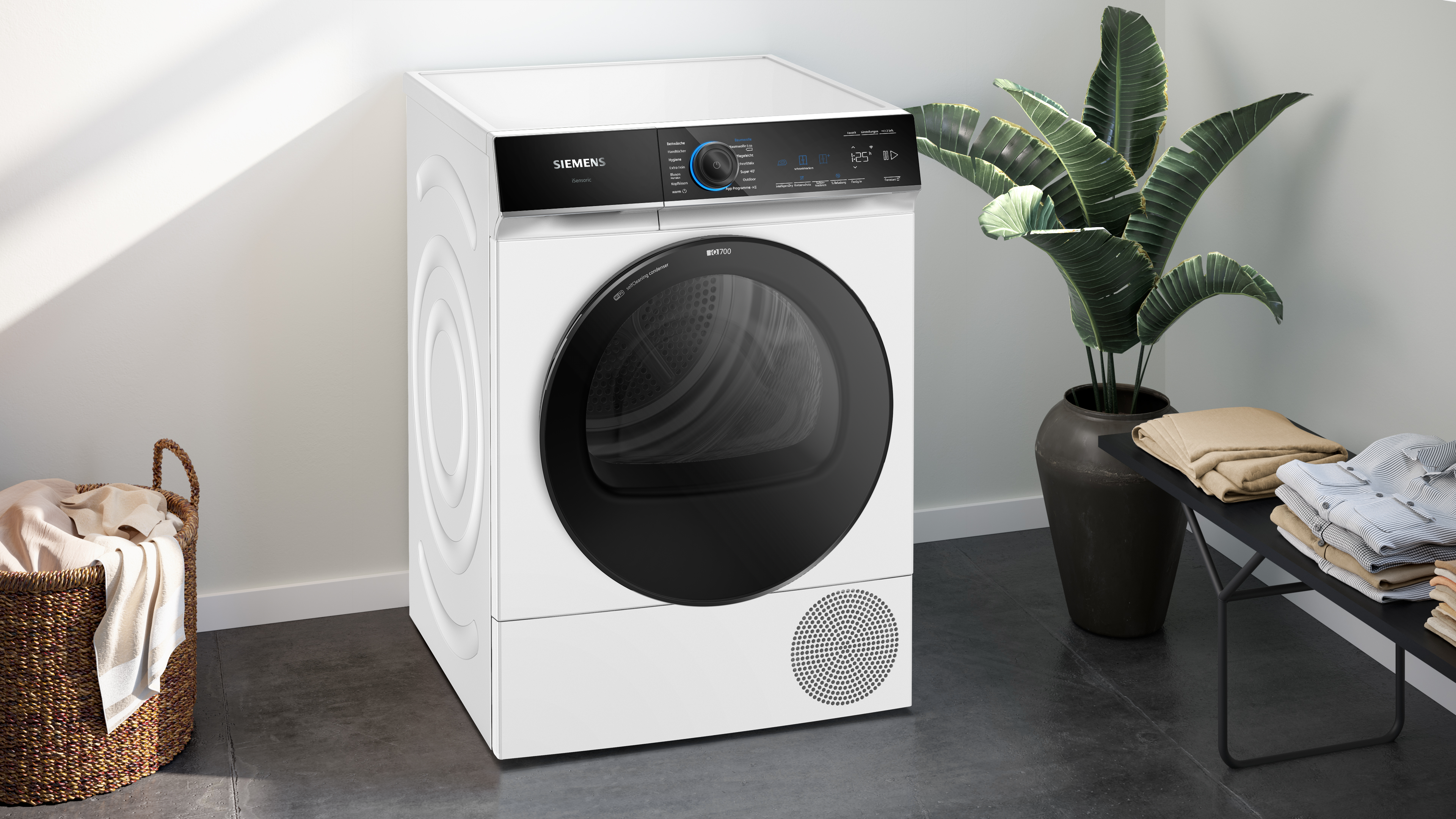 Weißer Siemens Trockner mit Digitalanzeige, Korb und Pflanze. Beige Hemden liegen auf einer Bank.