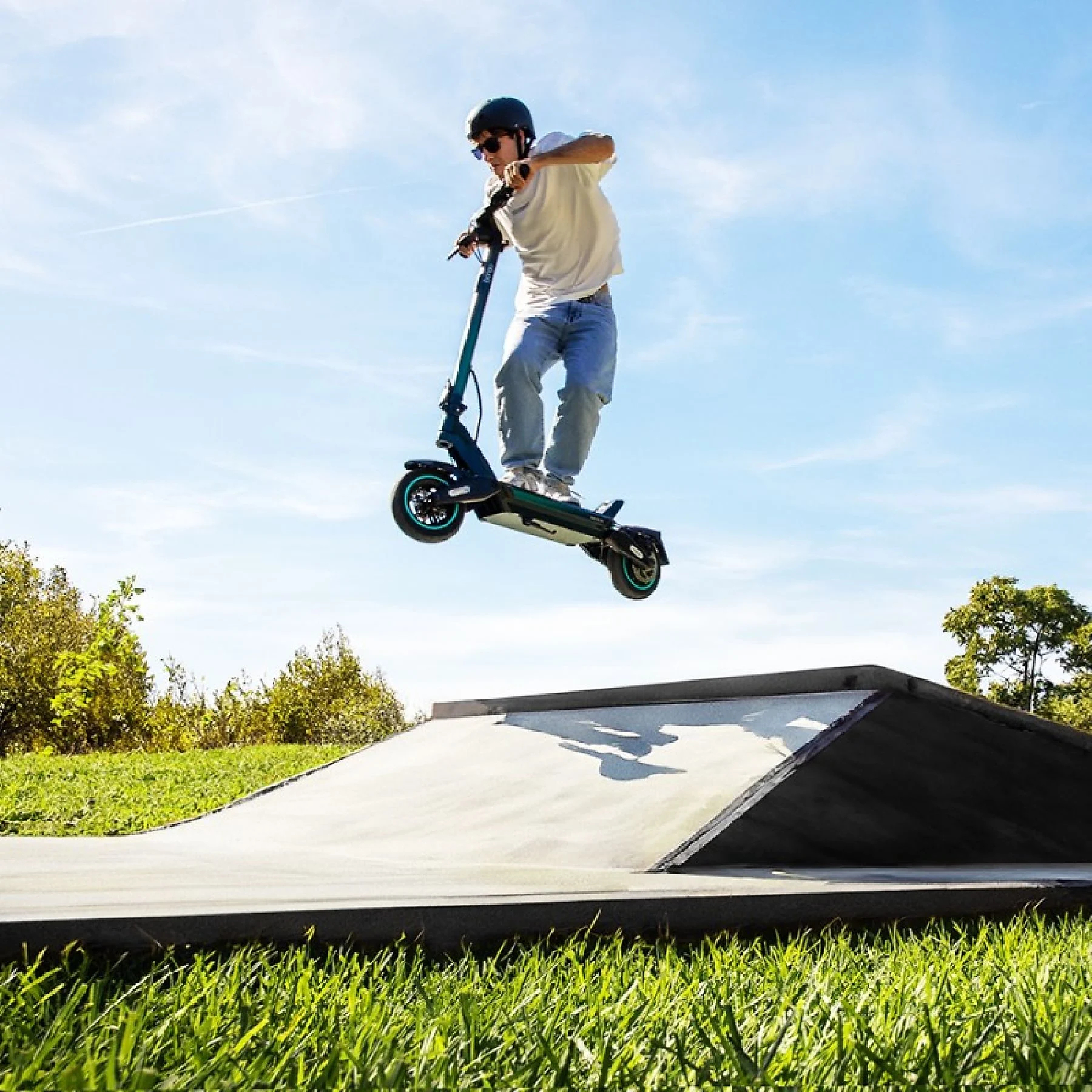 Mężczyzna na skuterze skacze w powietrzu. Niebieskie niebo, zielona trawa i rampa w zasięgu wzroku.
