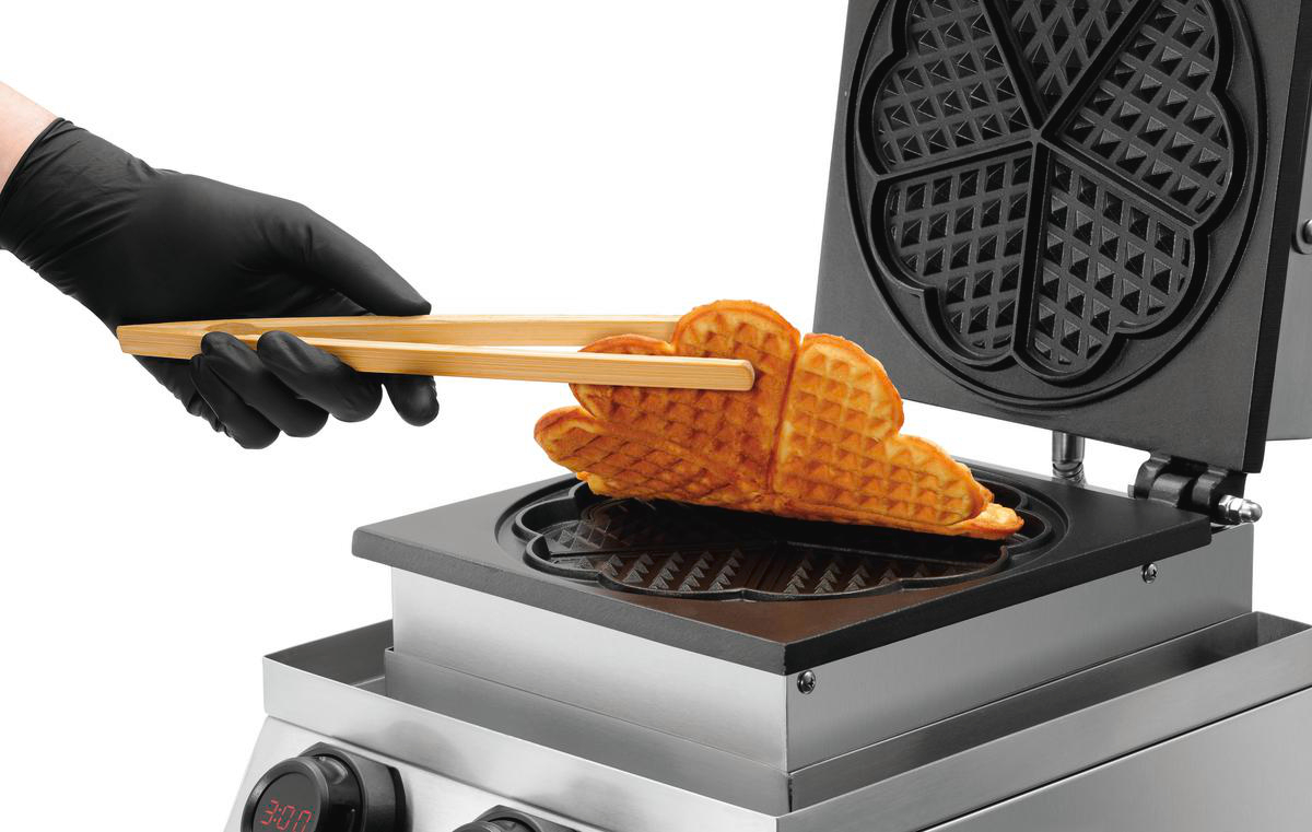 Eine Hand in schwarzer Handschuh benutzt Zange, um ein Herz-Waffel aus einer Waffelmaschine zu nehmen.