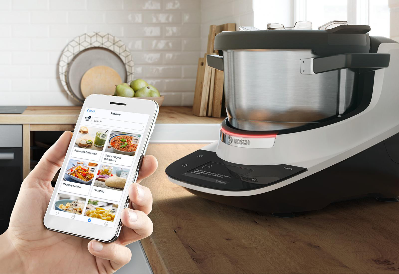 Eine Person hält ein Smartphone mit einer Rezeptliste, im Hintergrund eine Bosch Küchenmaschine.