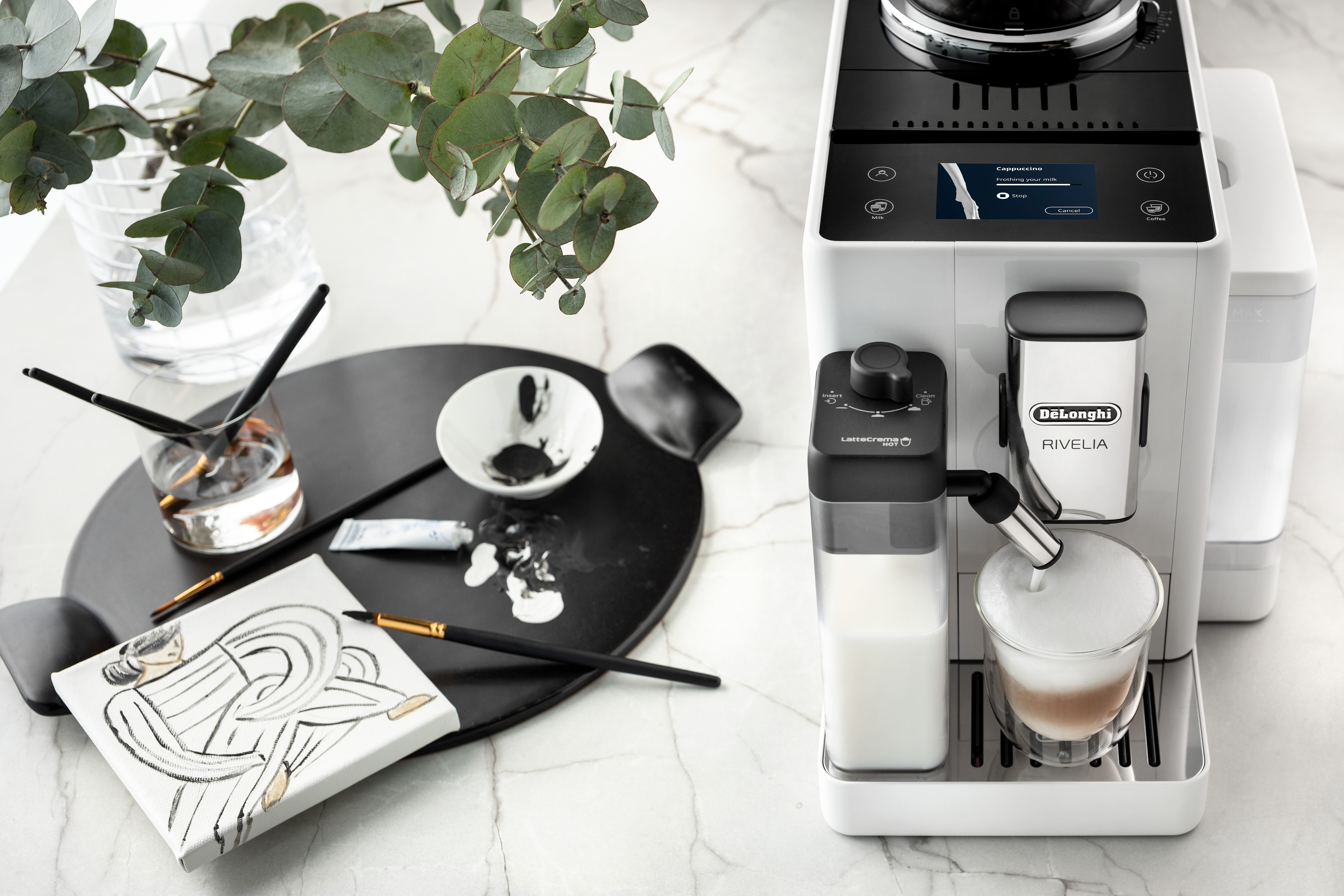 Weiß-schwarze Kaffeemaschine mit Milchaufschäumer und Malutensilien auf einem Tisch.