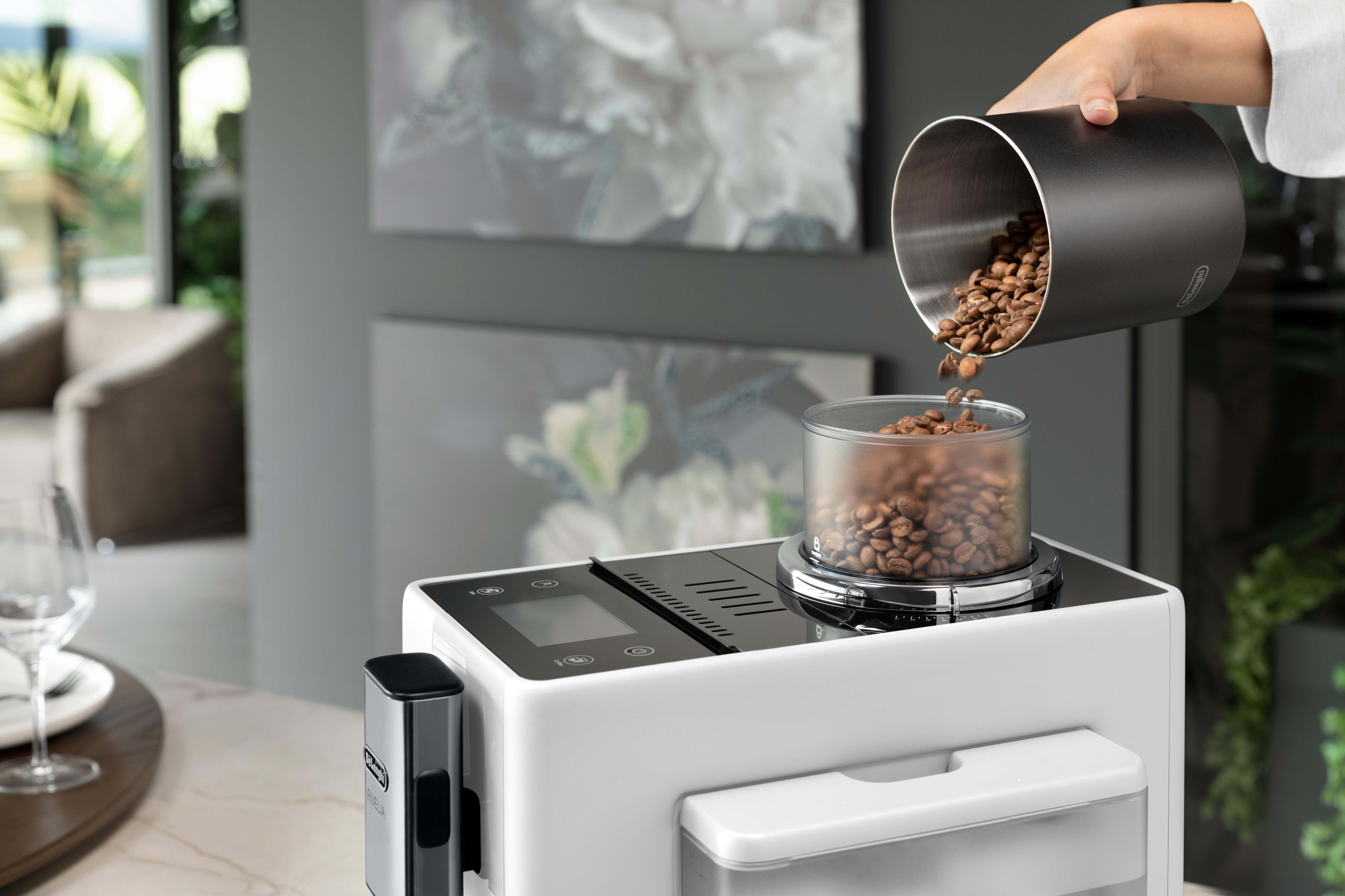 Eine Hand gießt Kaffeebohnen in eine Kaffeemaschine. Die Maschine ist weiß und schwarz.