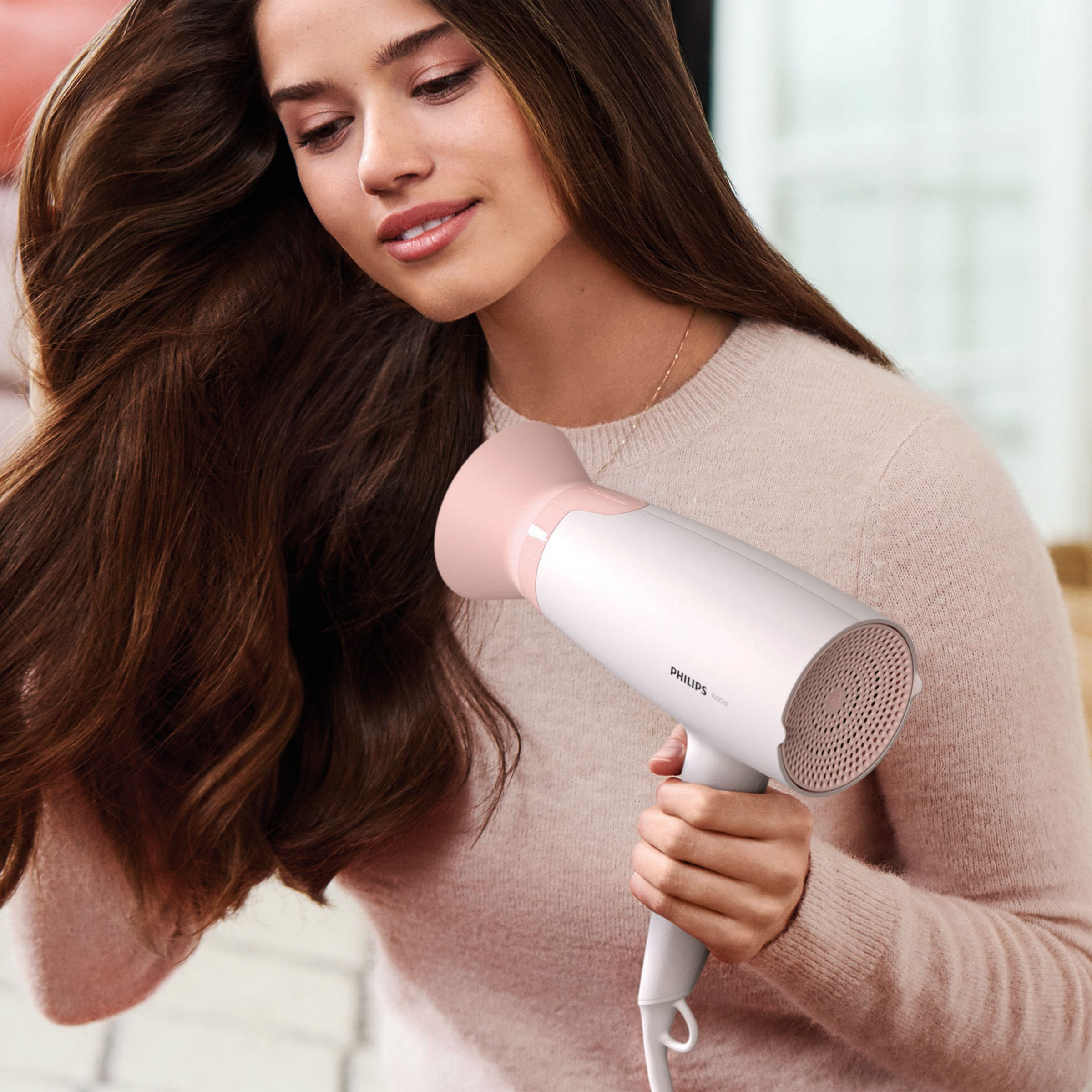Kobieta trzymająca różowo-białą suszarkę do włosów, susząca swoje długie brązowe włosy. Ma na sobie jasny sweter.