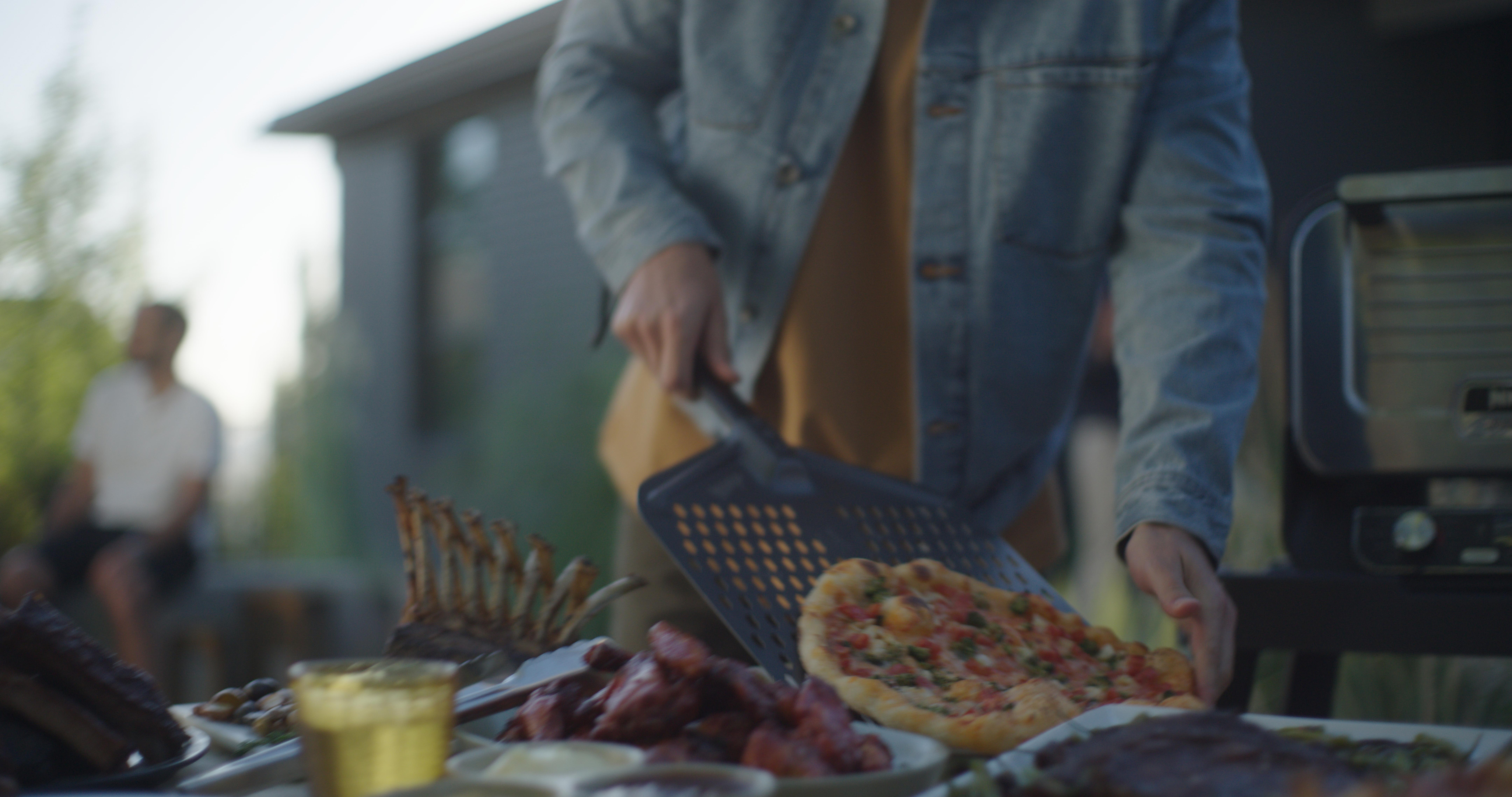 Person bereitet Essen vor, inkl. Pizza, auf einem Tisch im Freien. Eine weitere Person sitzt.
