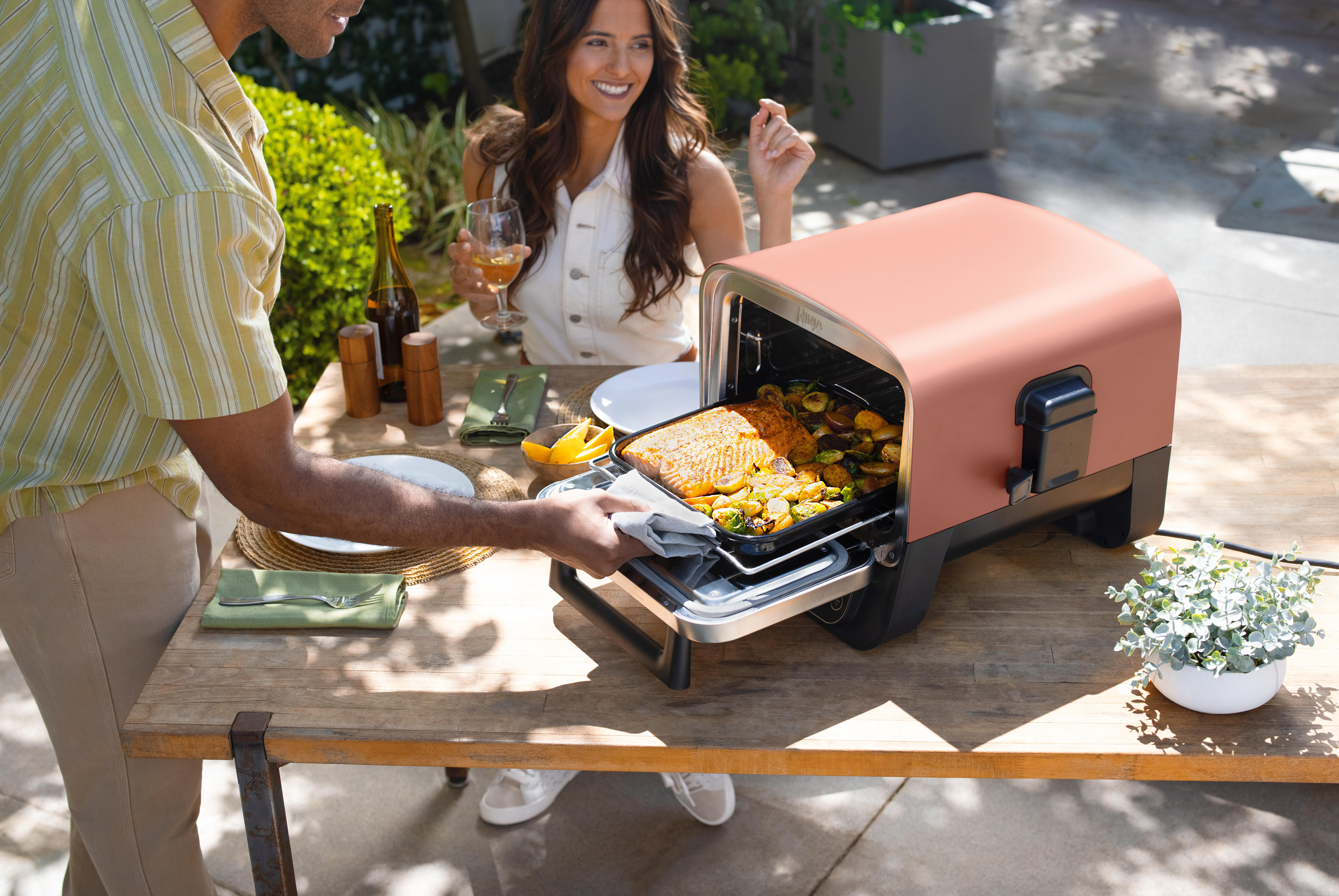 Ein Paar bereitet Essen mit einem rosa Tischplattenofen zu. Das Essen befindet sich in einem Tablett. Die Szene ist draußen.