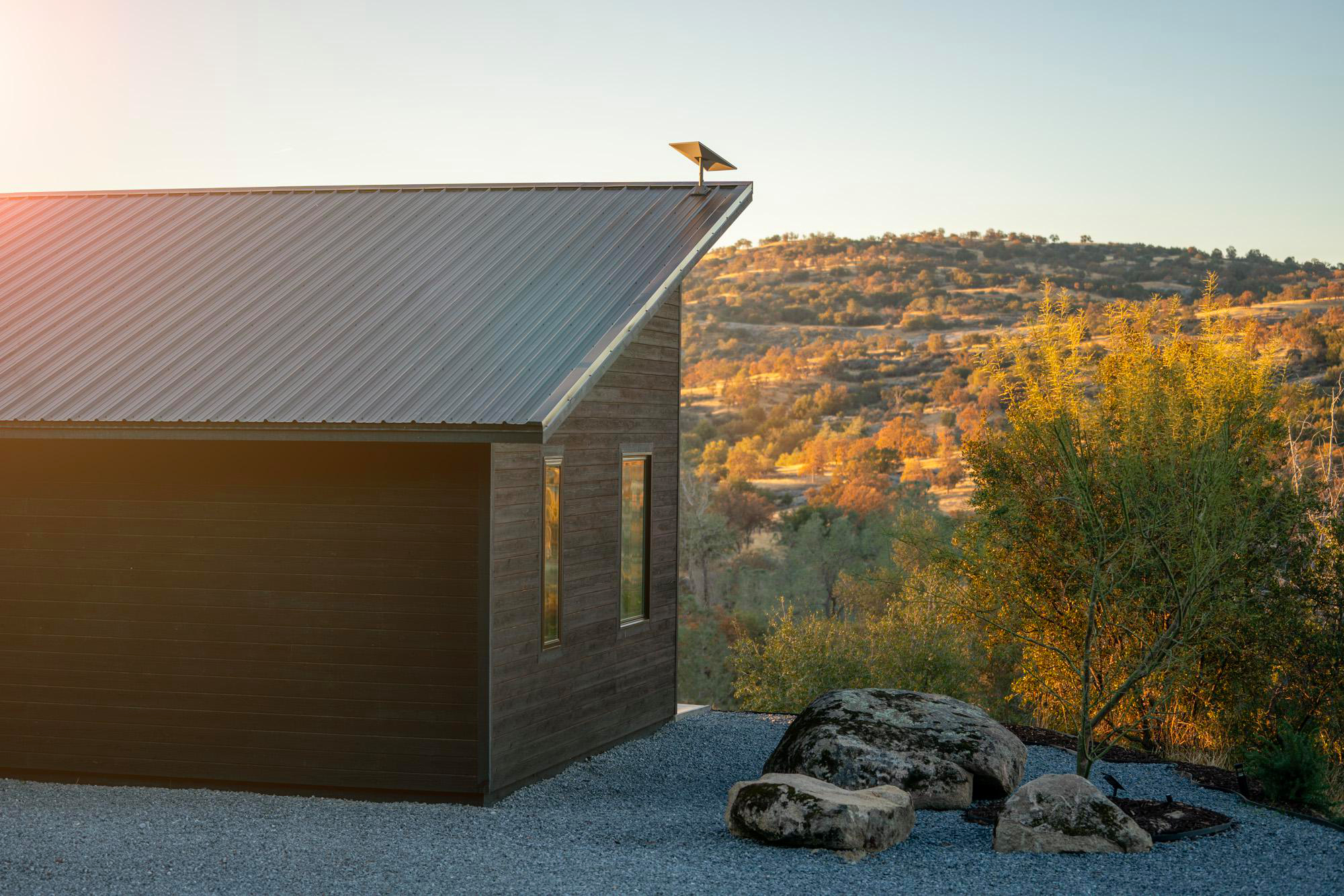 Haus mit dunkler Verkleidung und Metalldach, Felsen im Vordergrund. Sonnenuntergang.
