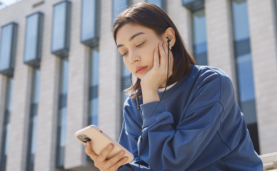 Una donna seduta fuori ascolta musica sul suo telefono con gli auricolari, guardando in basso.