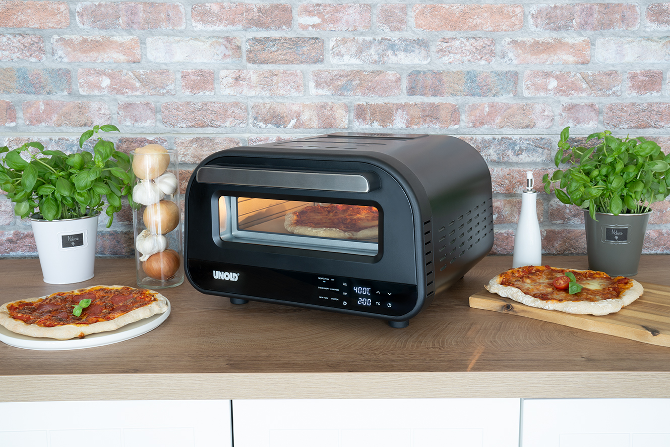 Schwarzer Pizzaofen mit Pizza, weitere Pizzen seitlich, Pflanzen.