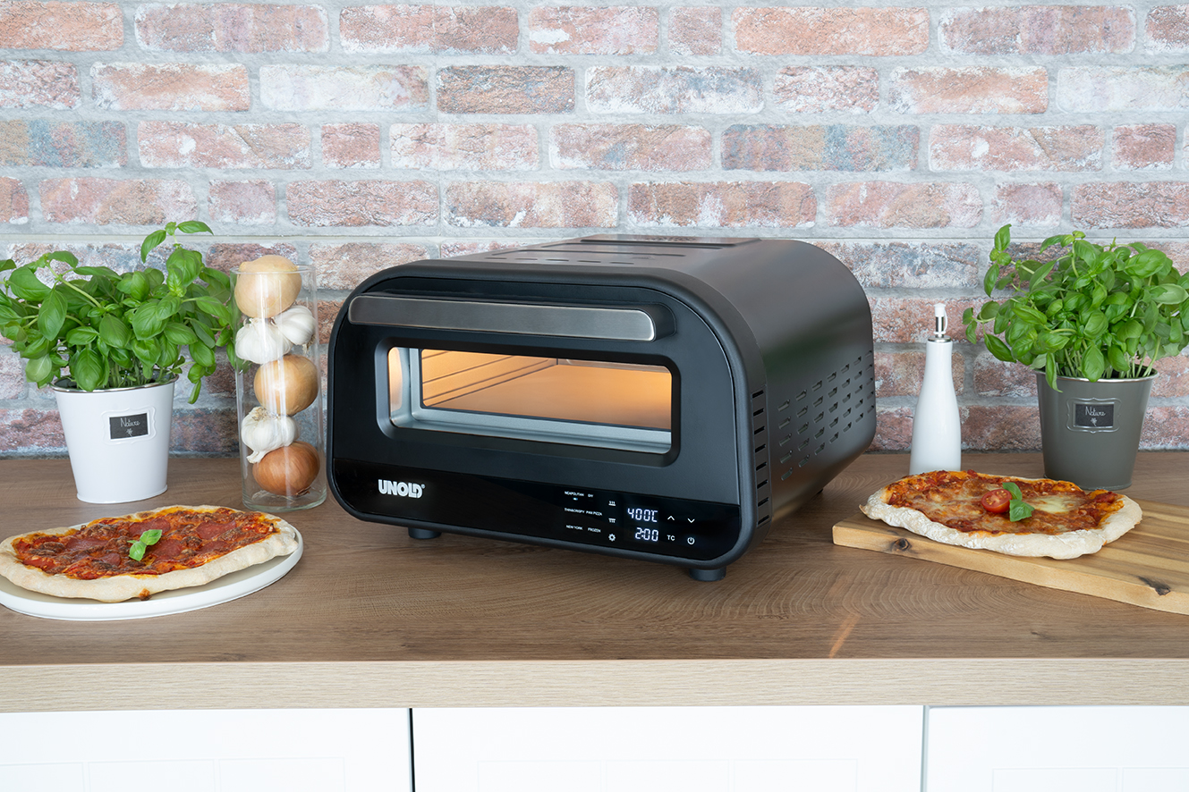 Schwarzer Pizzaofen mit zwei Pizzen, Basilikum und Zwiebeln auf einem Holztisch.