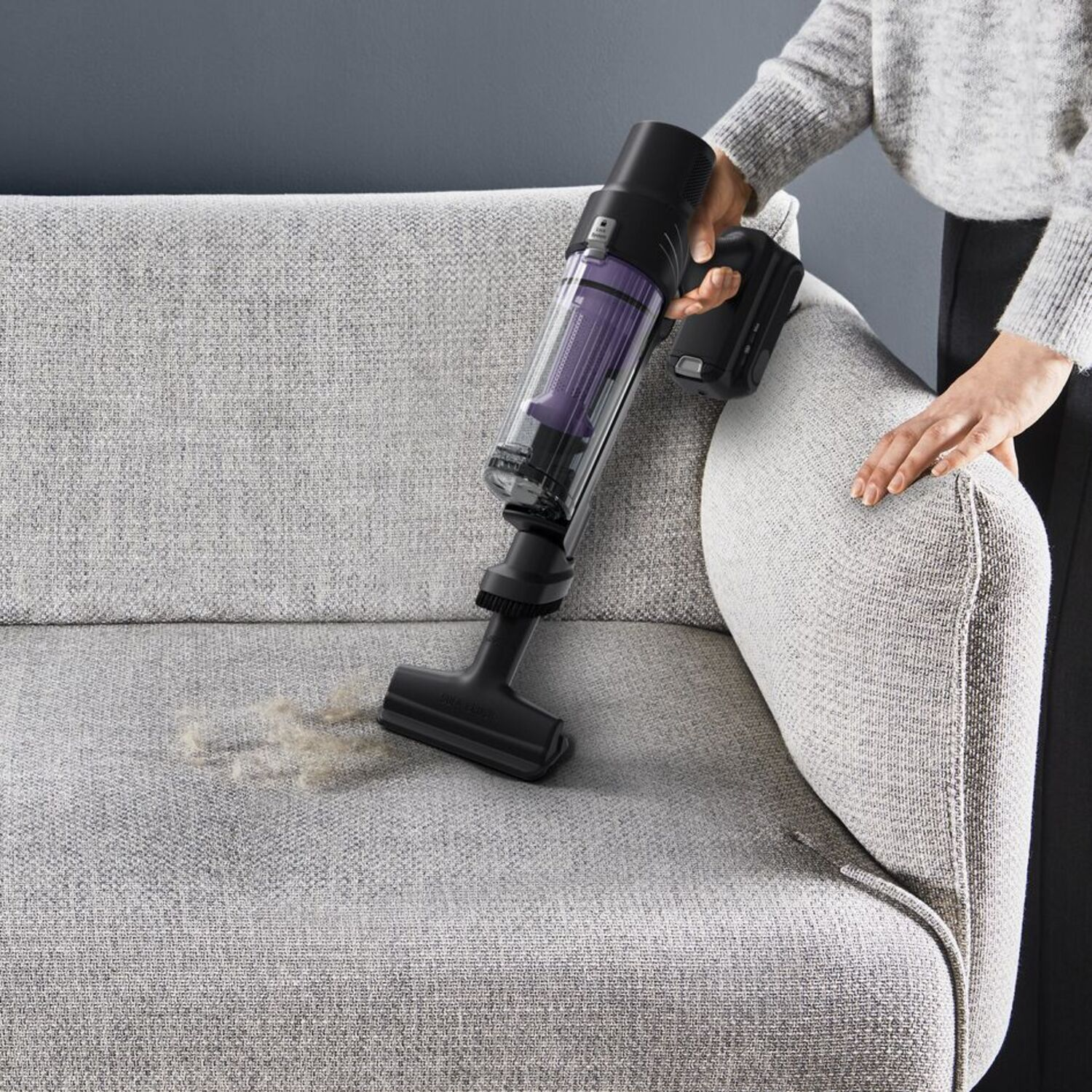 A person vacuums a gray couch with a cordless vacuum cleaner. Dust and debris are visible on the couch.