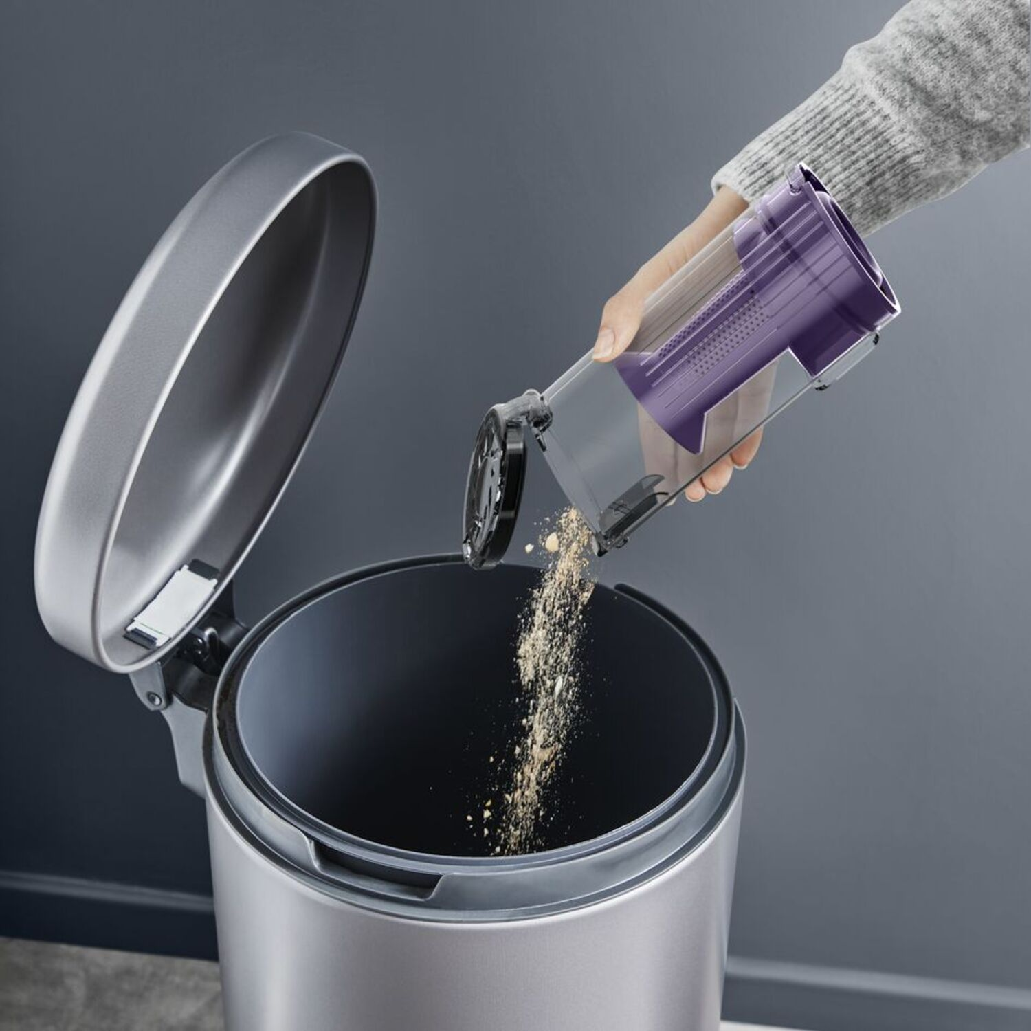 A hand emptying a vacuum cleaner dustbin into a trash can. Grey and purple colors.