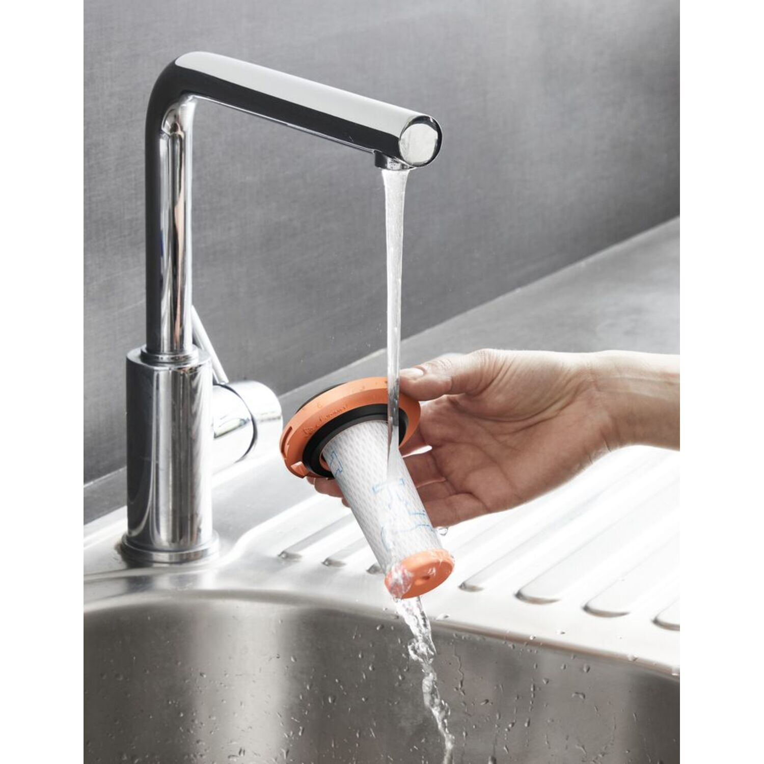 A hand rinsing a filter under a faucet. The filter is cylindrical and has an orange seal.