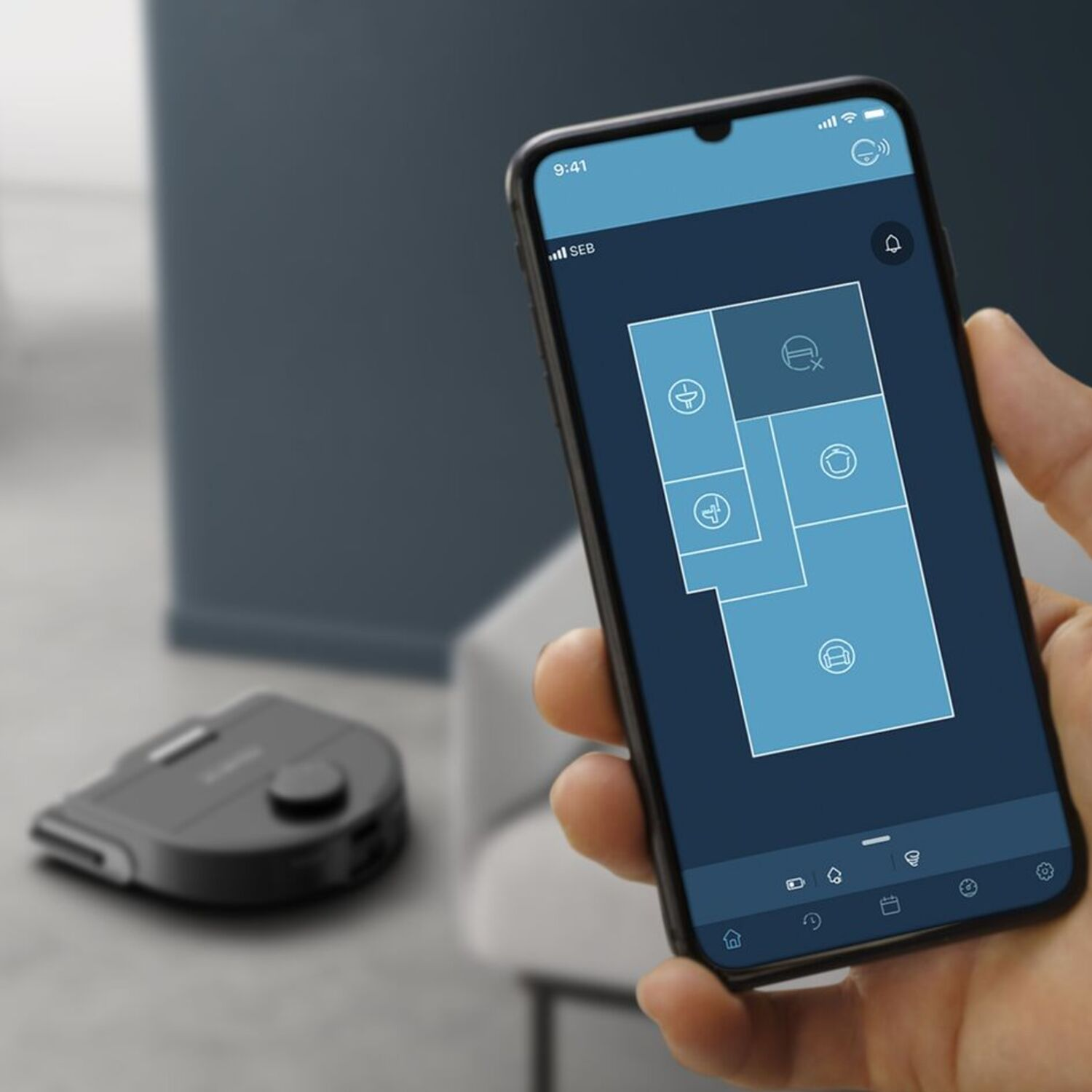 Hand holding a smartphone displaying a room plan with a cleaning robot in the background.