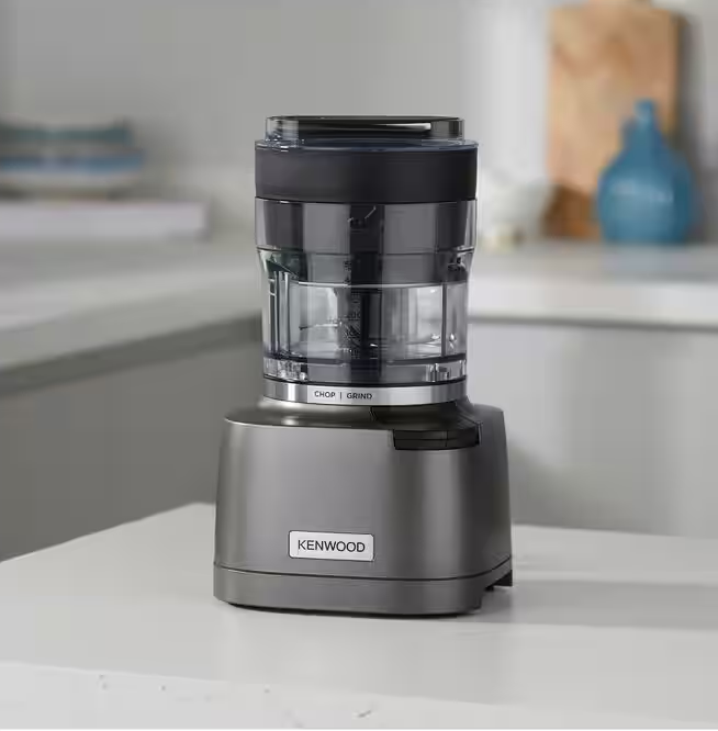 Kenwood-Zerkleinerer auf weißer Oberfläche. Graue Basis, schwarzer Deckel, durchsichtiger Behälter.