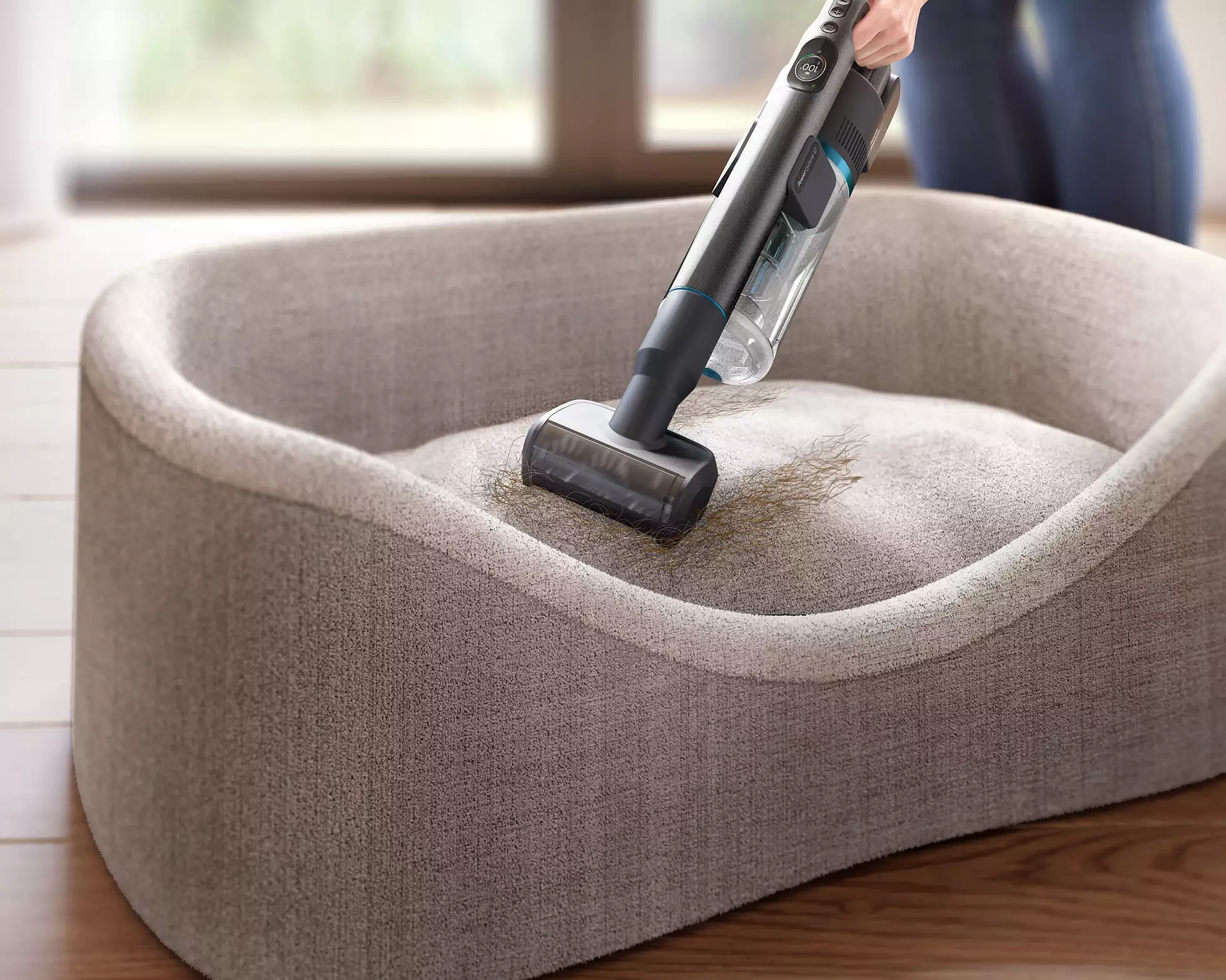 Una persona che aspira un letto per cani con un aspirapolvere portatile. Il letto è grigio e l'aspirapolvere è nero.