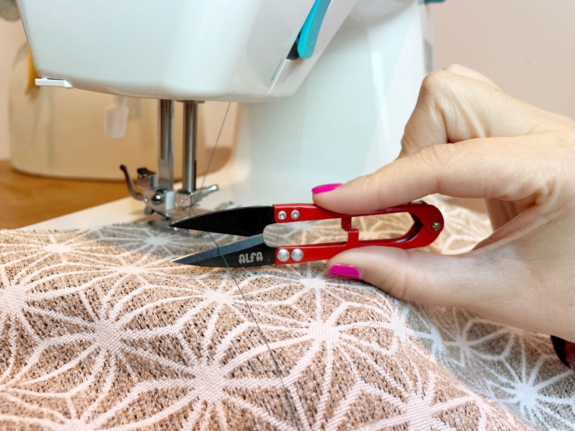 Una mano sostiene unas tijeras rojas pequeñas, cortando hilo cerca de una máquina de coser y tela.