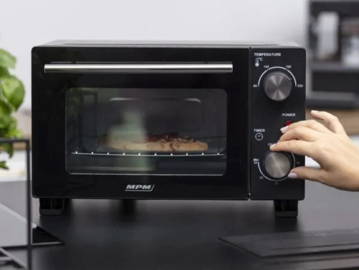 Czarny piekarnik na blacie. Ręka przekręca pokrętło. W środku pizza.