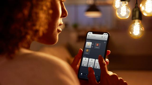 Frau mit Smartphone steuert Smart-Leuchten. Hintergrund zeigt hängende Glühbirnen mit warmer Beleuchtung.