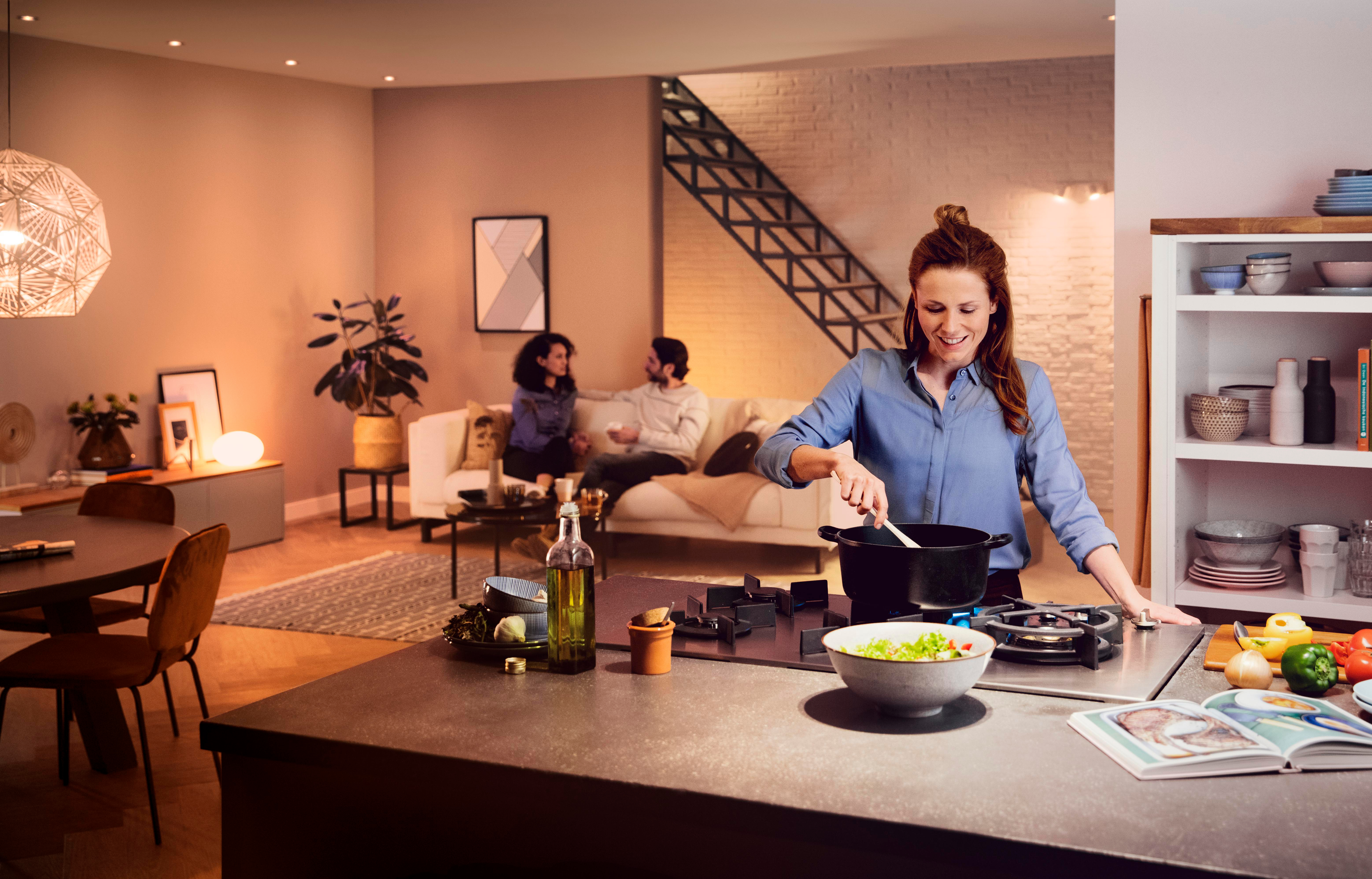 Eine Frau kocht in einer Küche. Zwei Personen sitzen im Hintergrund auf einem Sofa.