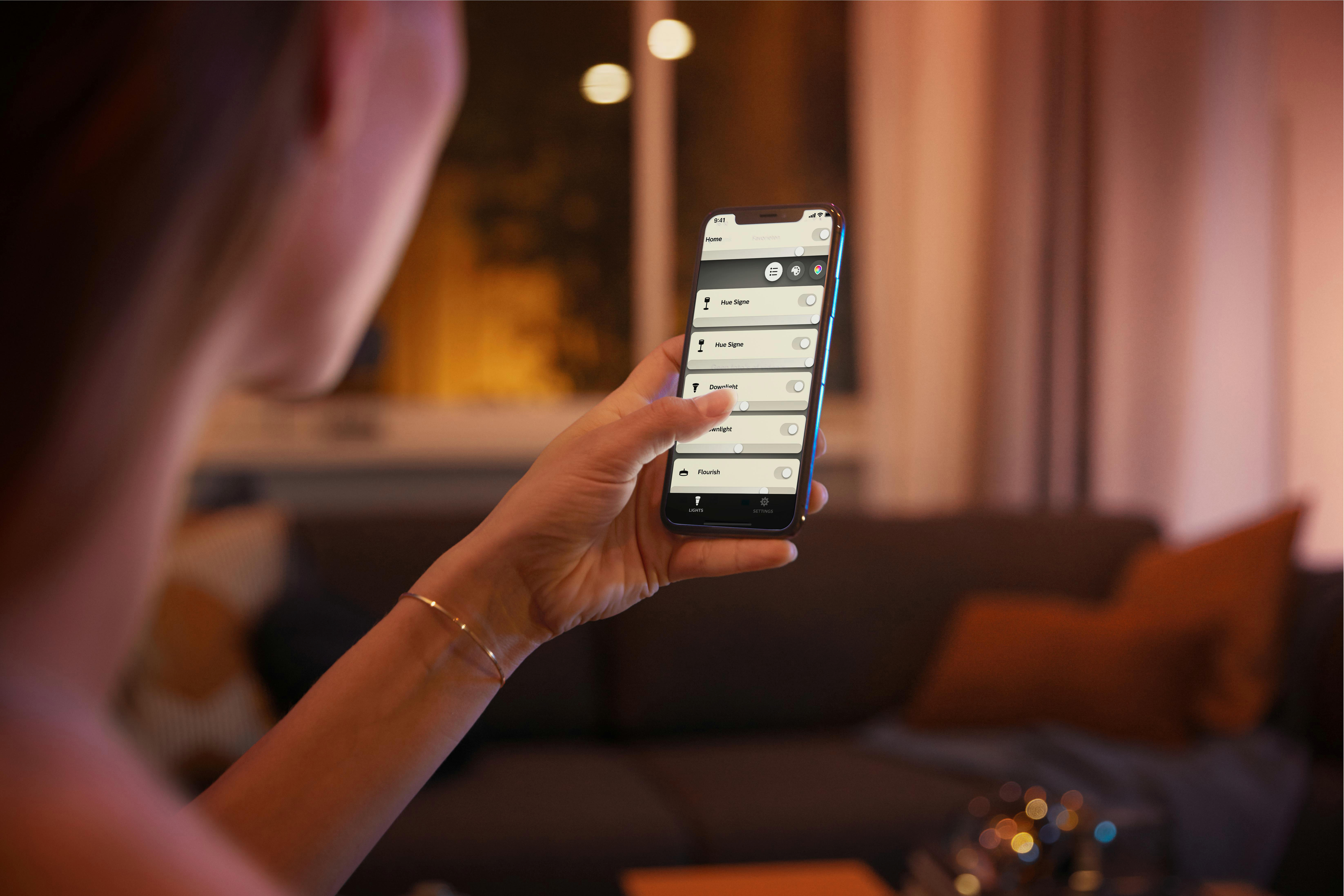 Frau hält Smartphone mit App-Oberfläche. Im Hintergrund ein Wohnzimmer mit Sofa.