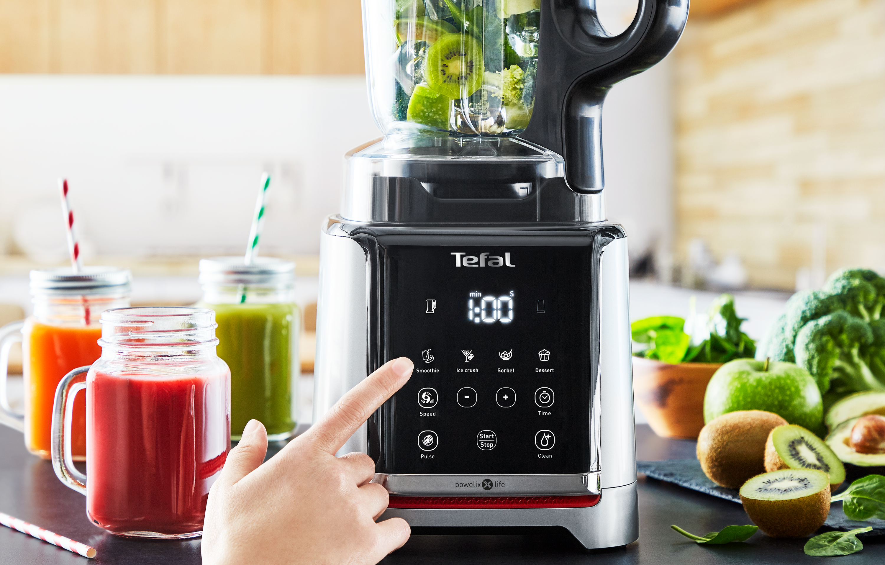 Ein Mixer mit Obst und eine Hand, die Knöpfe drückt, mit Smoothie-Gläsern im Hintergrund.