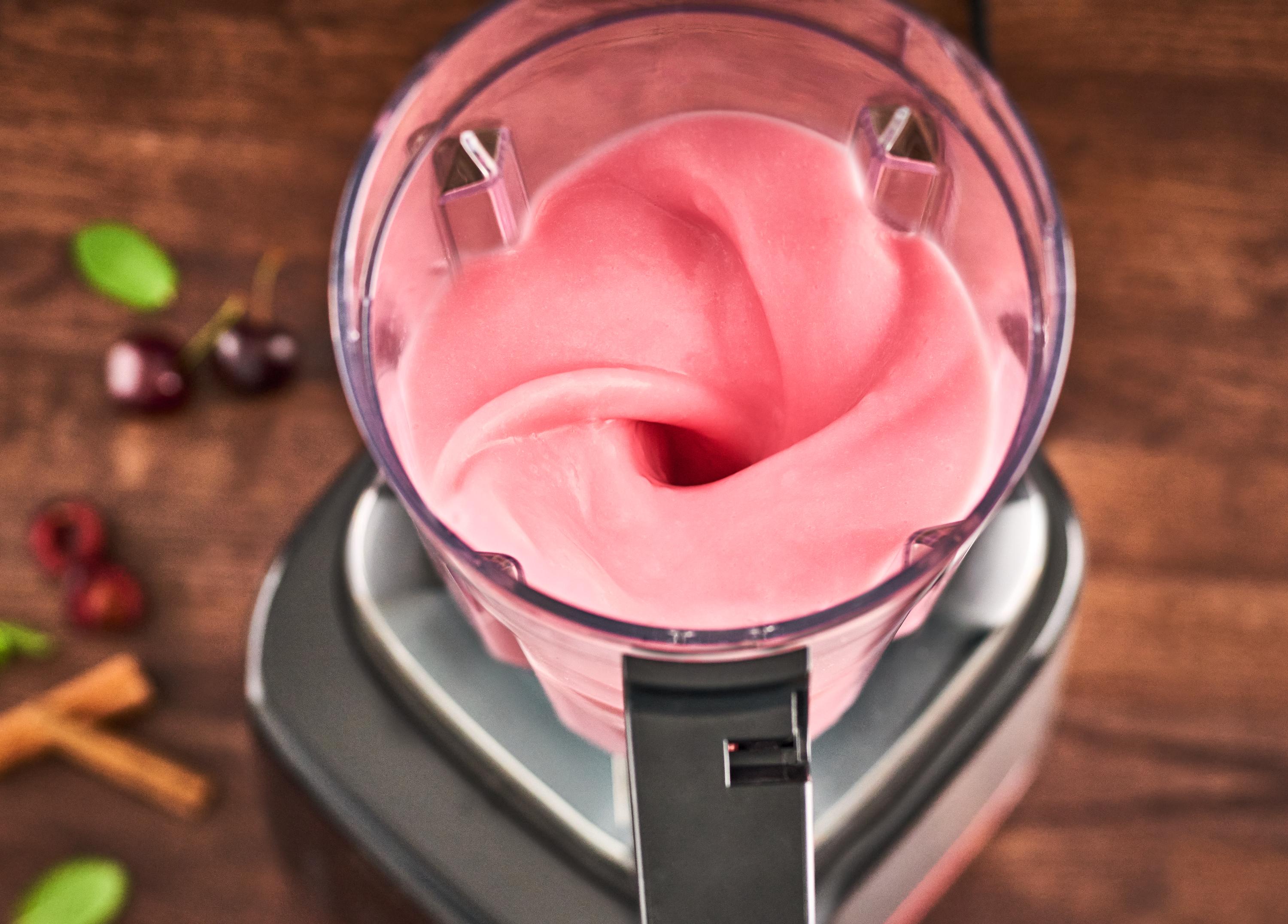 Mixer mit rosa Smoothie, von oben betrachtet, auf Holzoberfläche, mit Kirschen und Zimt.