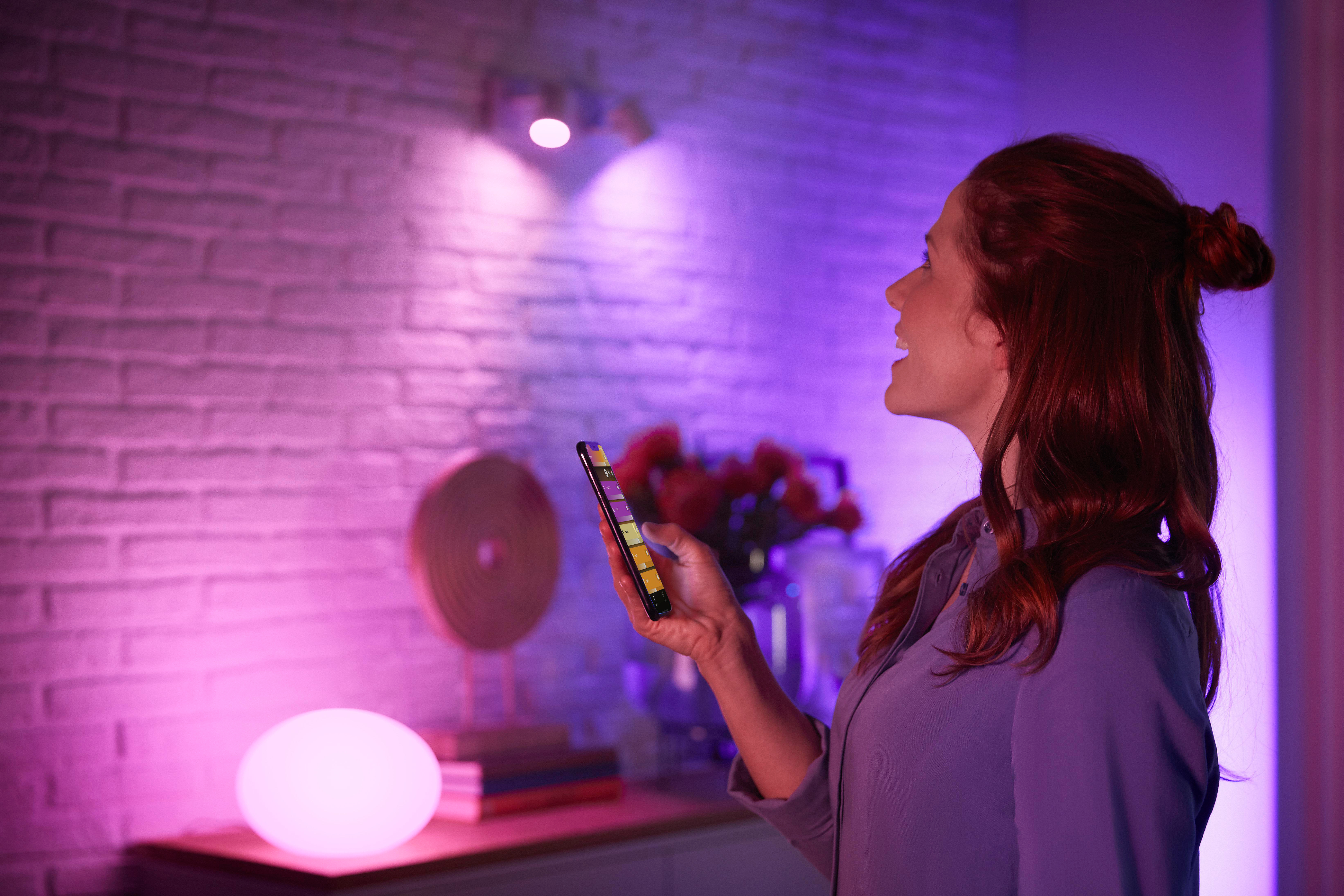 Frau mit Telefon, Lichtern und Lampe vor einer Ziegelmauer. Rosa- und Lilatöne erhellen den Raum.