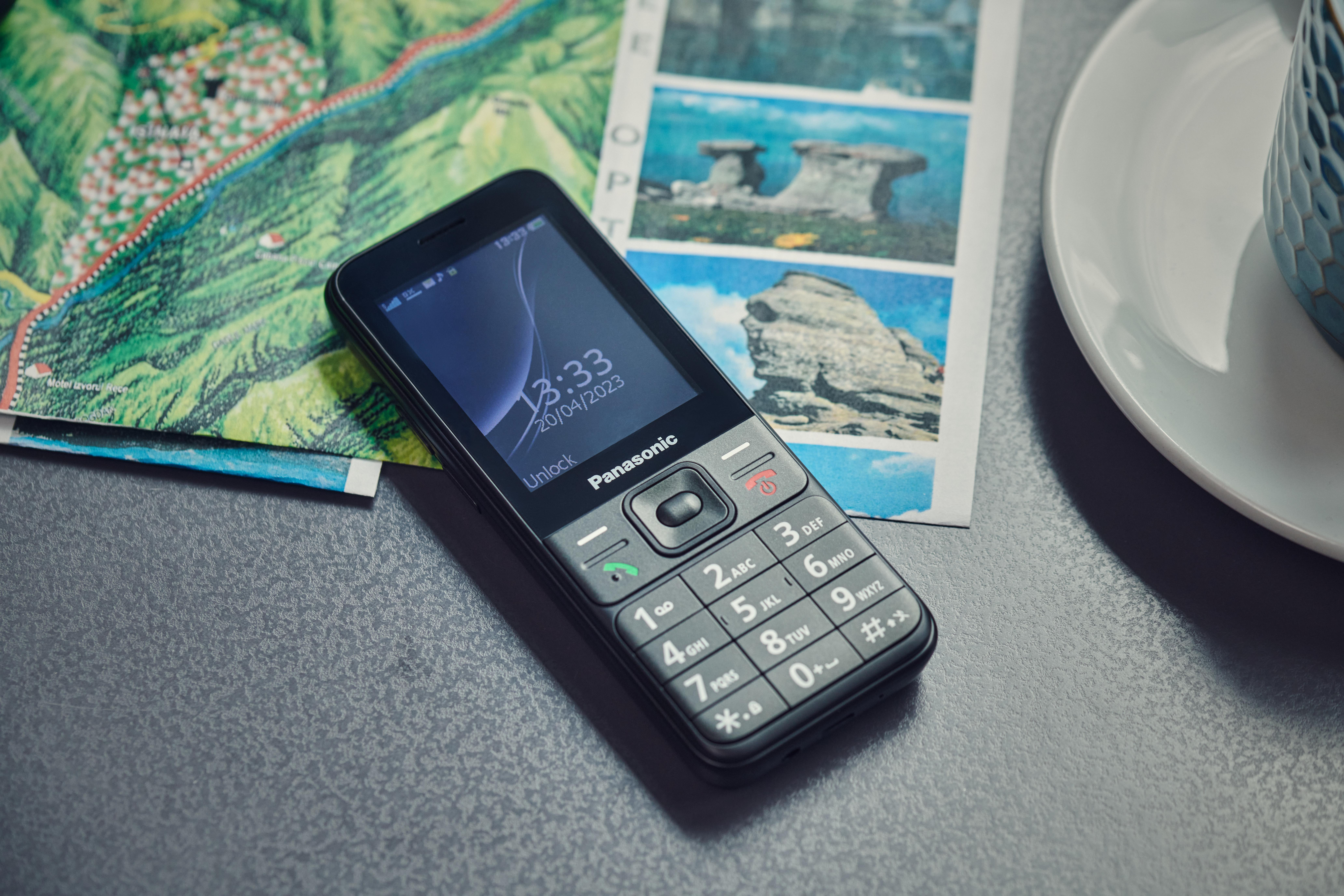 Schwarzes Panasonic-Telefon auf einem Tisch mit einer Karte und einer Tasse Kaffee. Das Telefon zeigt Zeit und Datum.