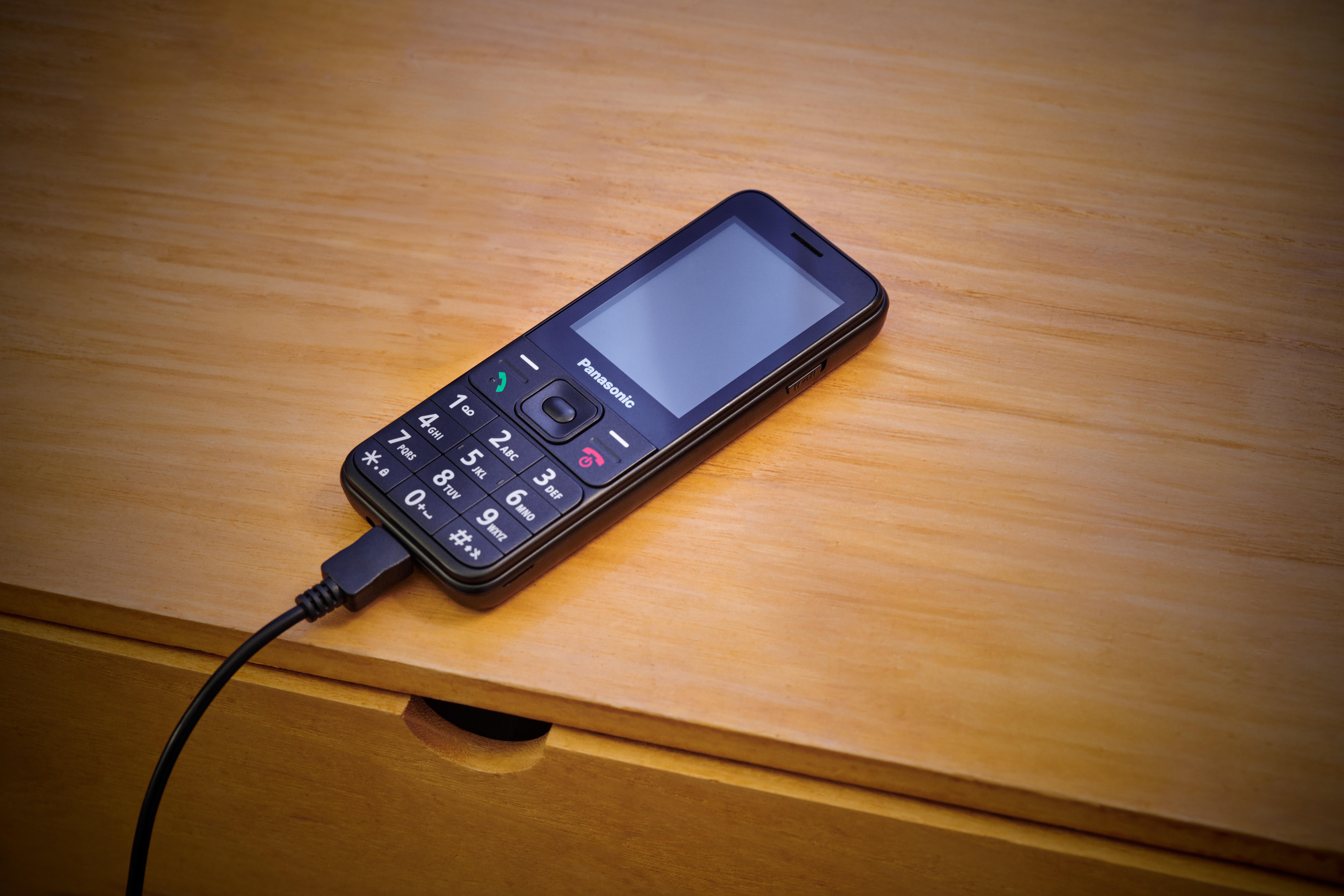Schwarzes Mobiltelefon, das auf einem Holztisch geladen wird. Das Telefon hat einen Bildschirm und Zifferntasten.