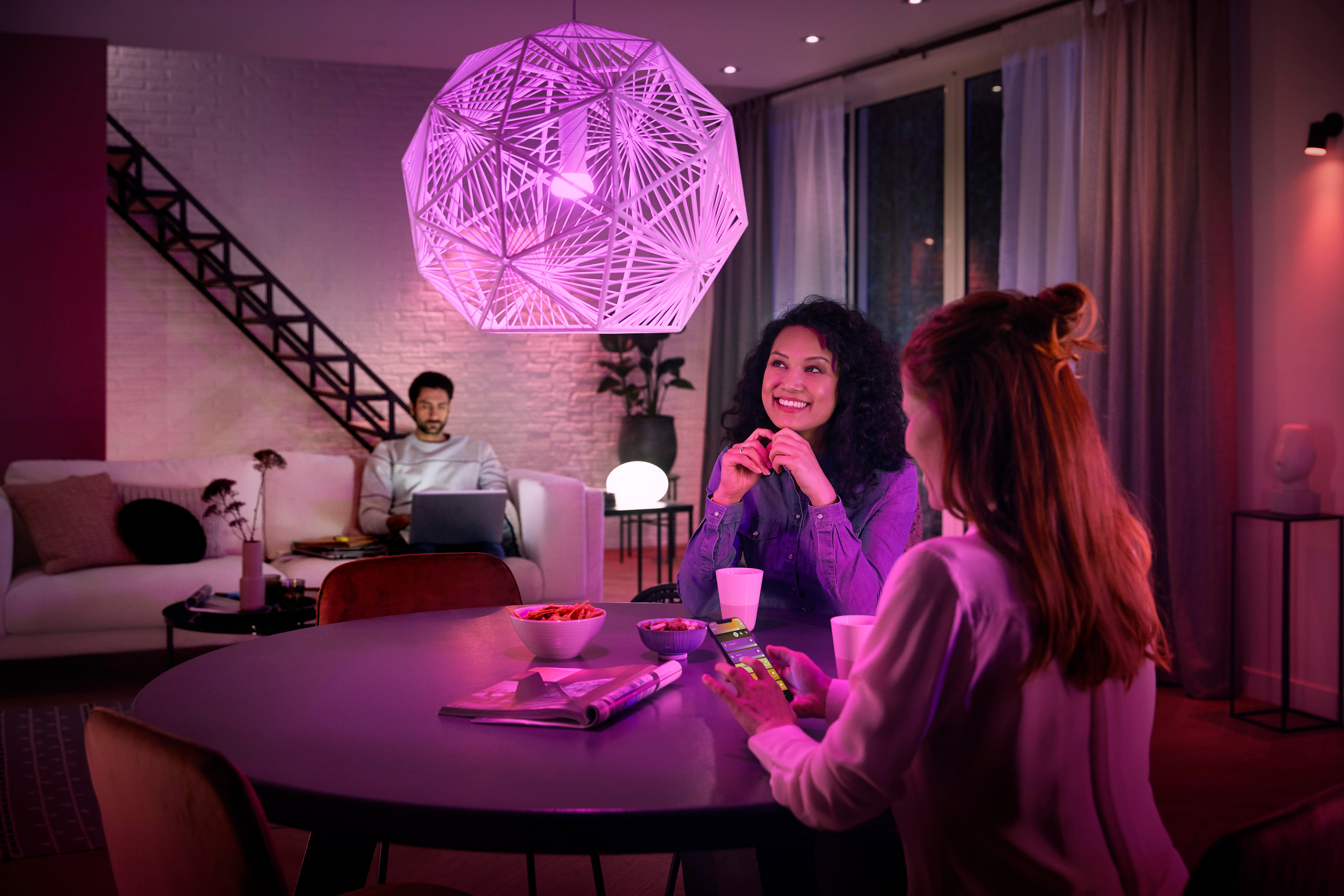 Personen sitzen an einem Tisch unter einem violetten Licht von einem geometrischen Kronleuchter. Ein Mann benutzt einen Laptop.