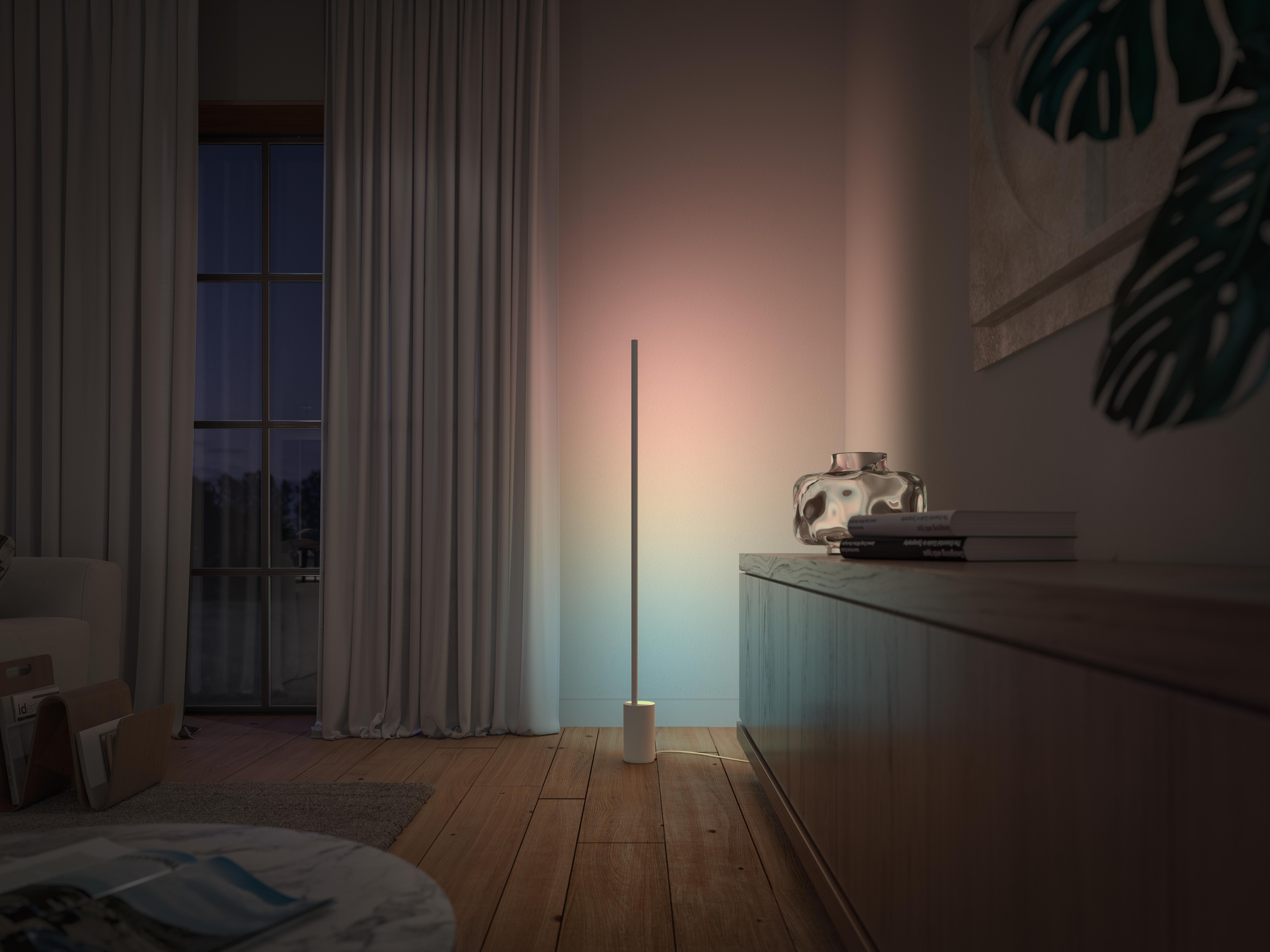 Eine hohe, farbenfrohe Lampe steht in einem Raum. Ein Fenster ist im Hintergrund. Ein Schrank und eine Vase stehen daneben.