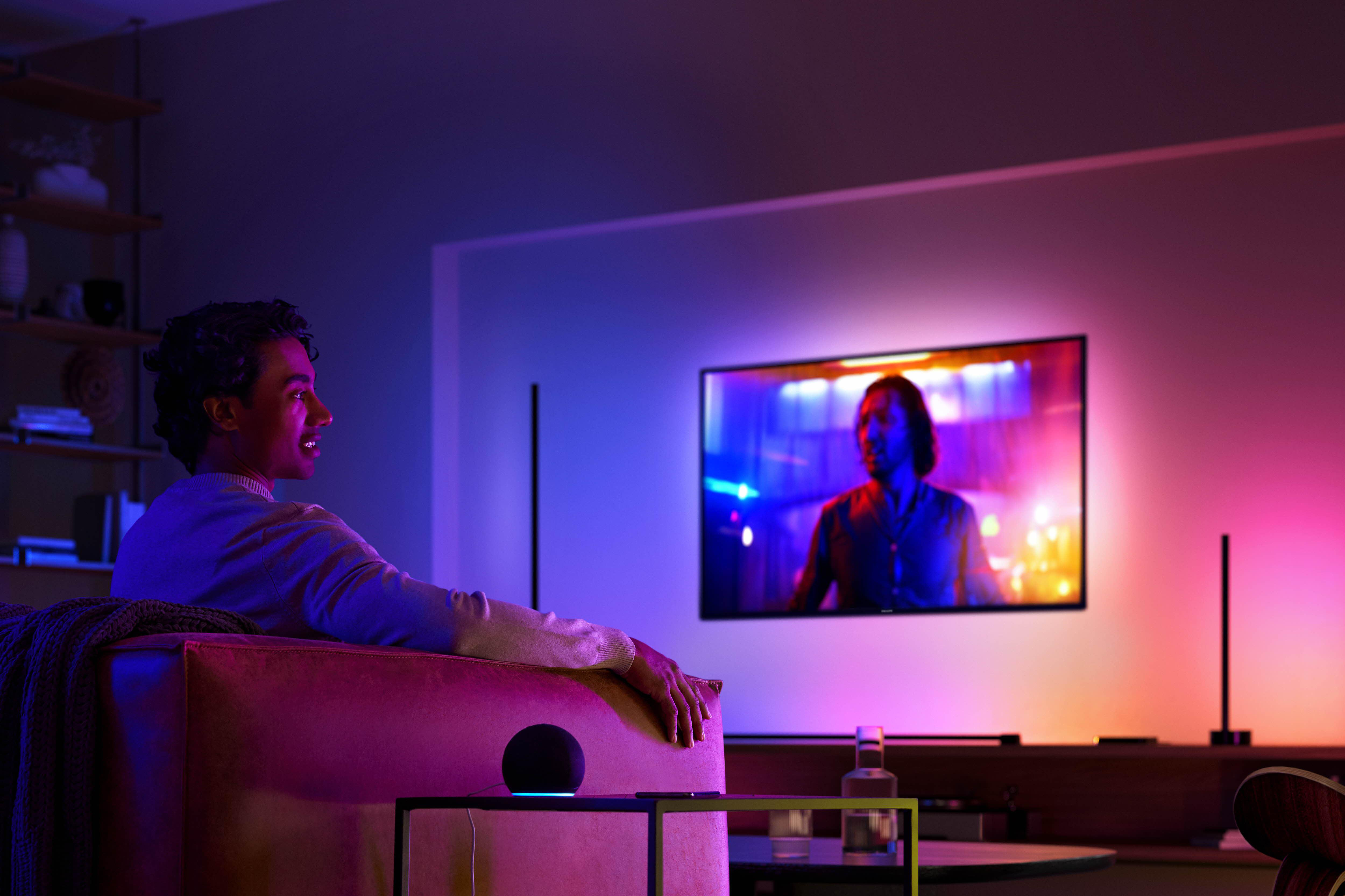 Person sieht im dunklen Raum fern. Der Fernseher zeigt eine Szene mit einem Mann. Alexa-Lautsprecher auf Tisch.