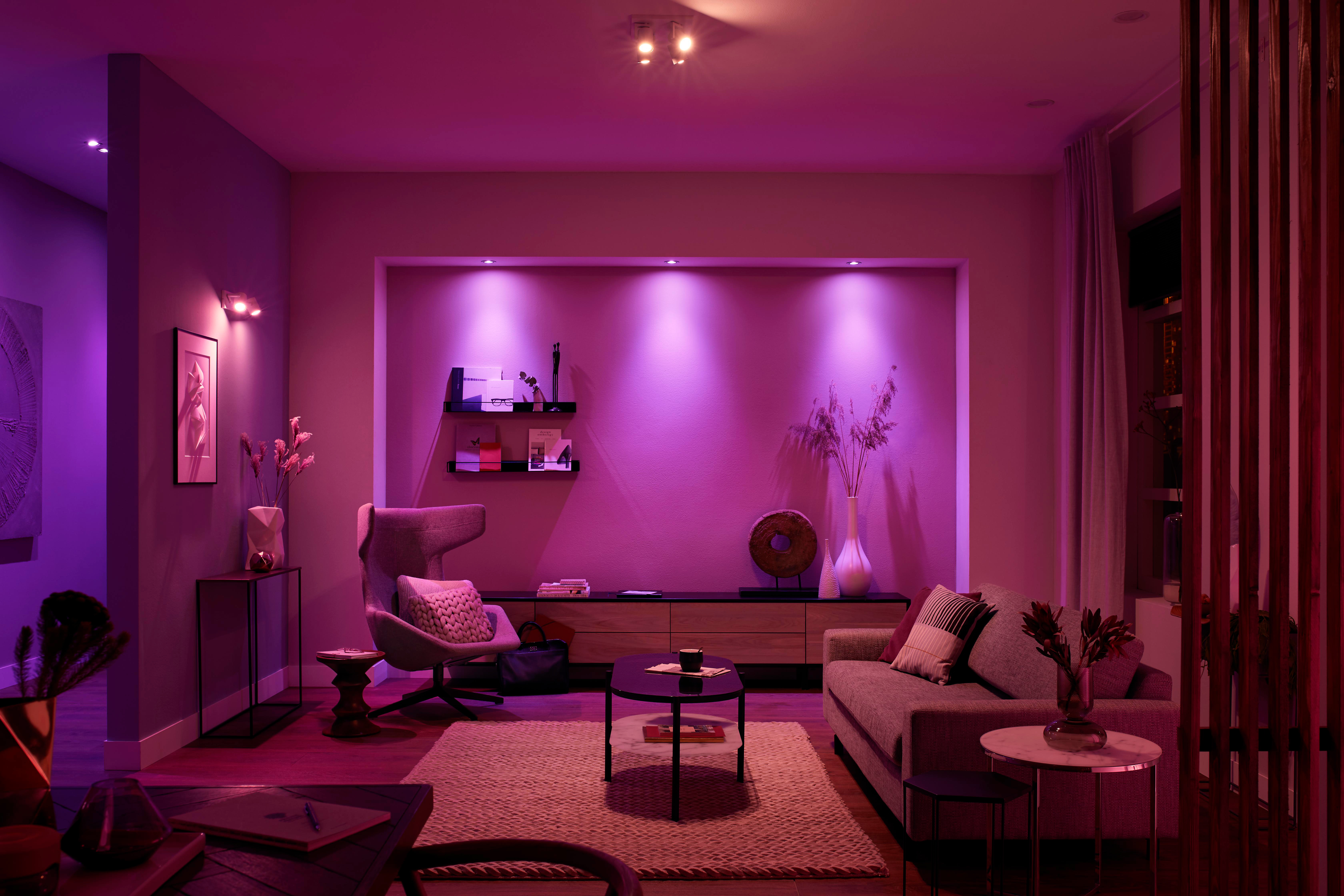 Ein Wohnzimmer mit lilafarbener Beleuchtung. Ein Sofa, ein Stuhl, ein Couchtisch und eine Holzverkleidung sind sichtbar. Ein Teppich liegt auf dem Boden.