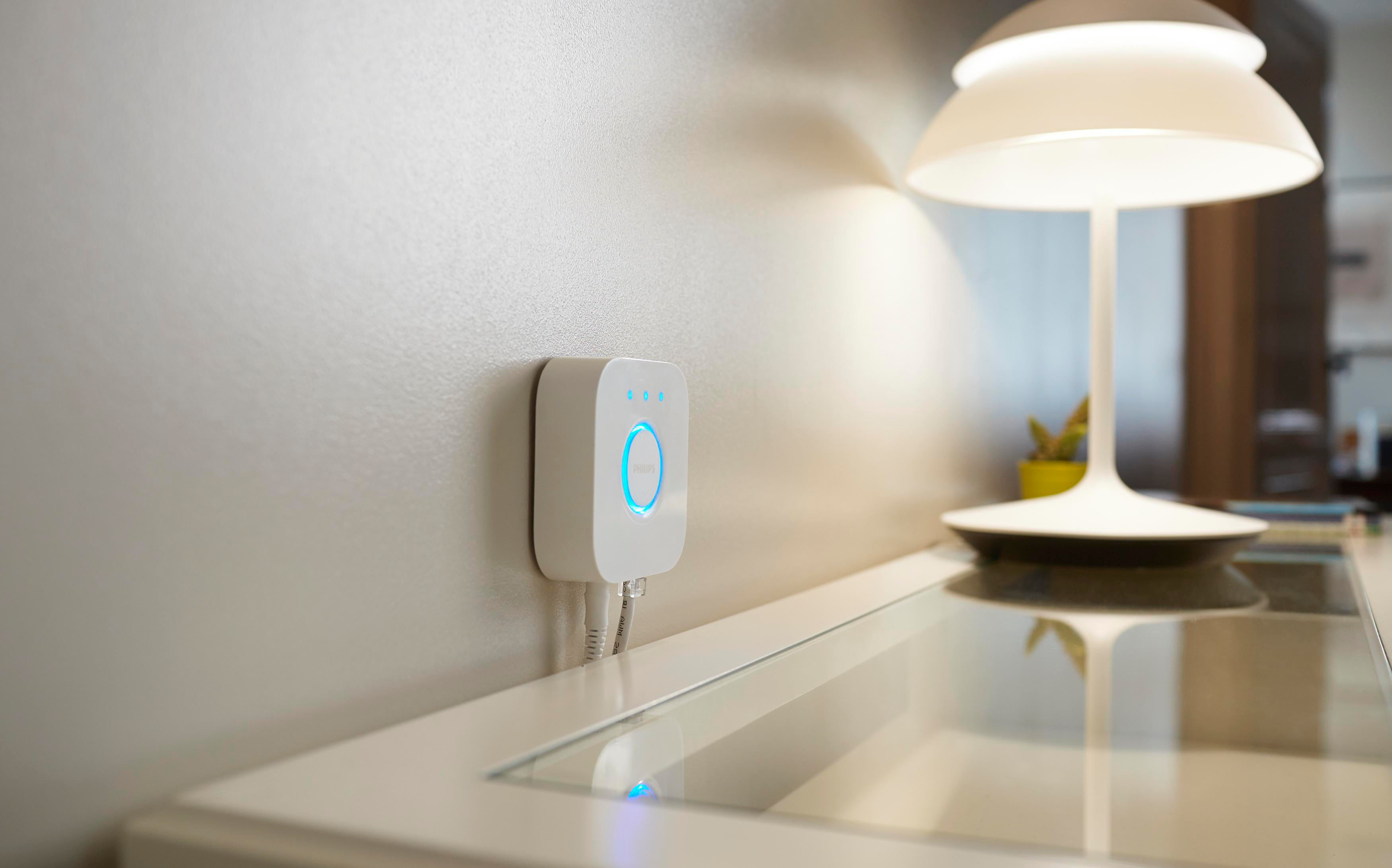 Ein weißer Smart-Home-Hub an einer Wand mit blauem Licht. Eine Lampe und eine Pflanze stehen auf einem Tisch.