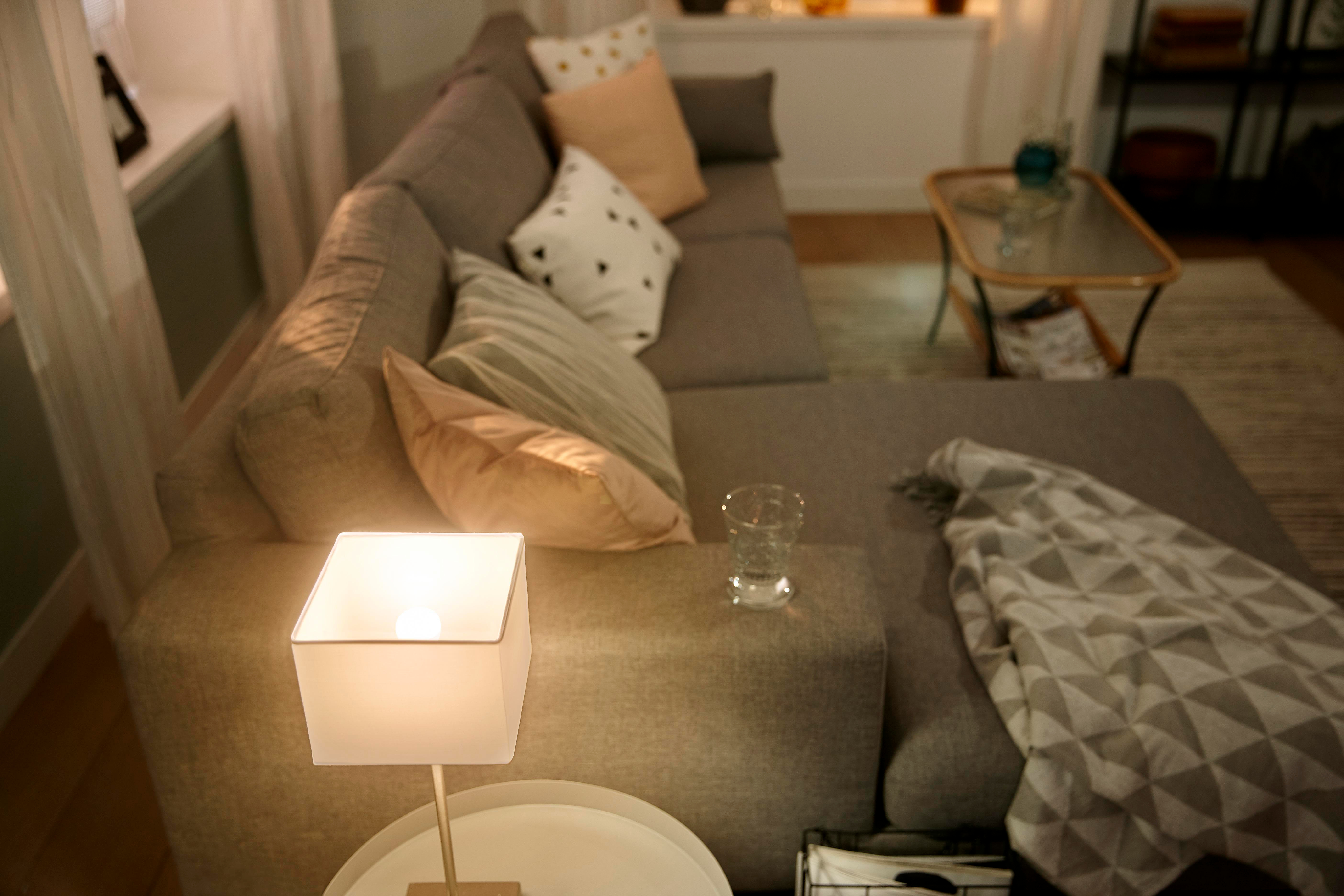Wohnzimmerszene mit grauem Sofa, Lampe, Glas und Kissen, die eine gemütliche Atmosphäre schaffen.