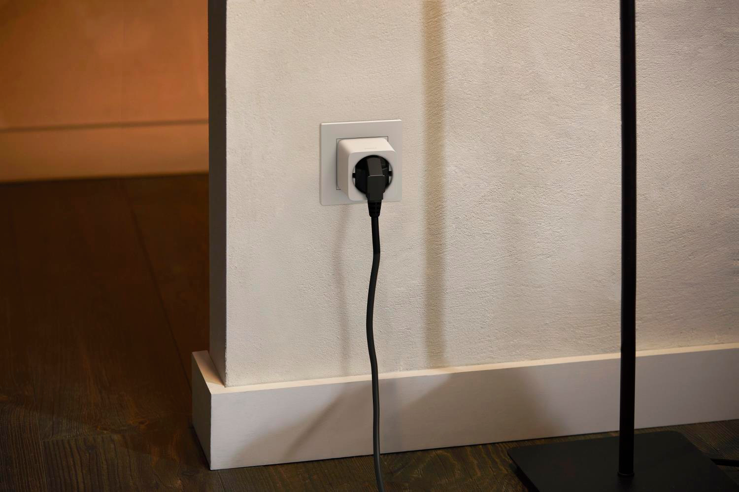 Ein schwarzer Stecker steckt in einer weißen Wandsteckdose an einer hellen Wand, mit Holzfußboden.